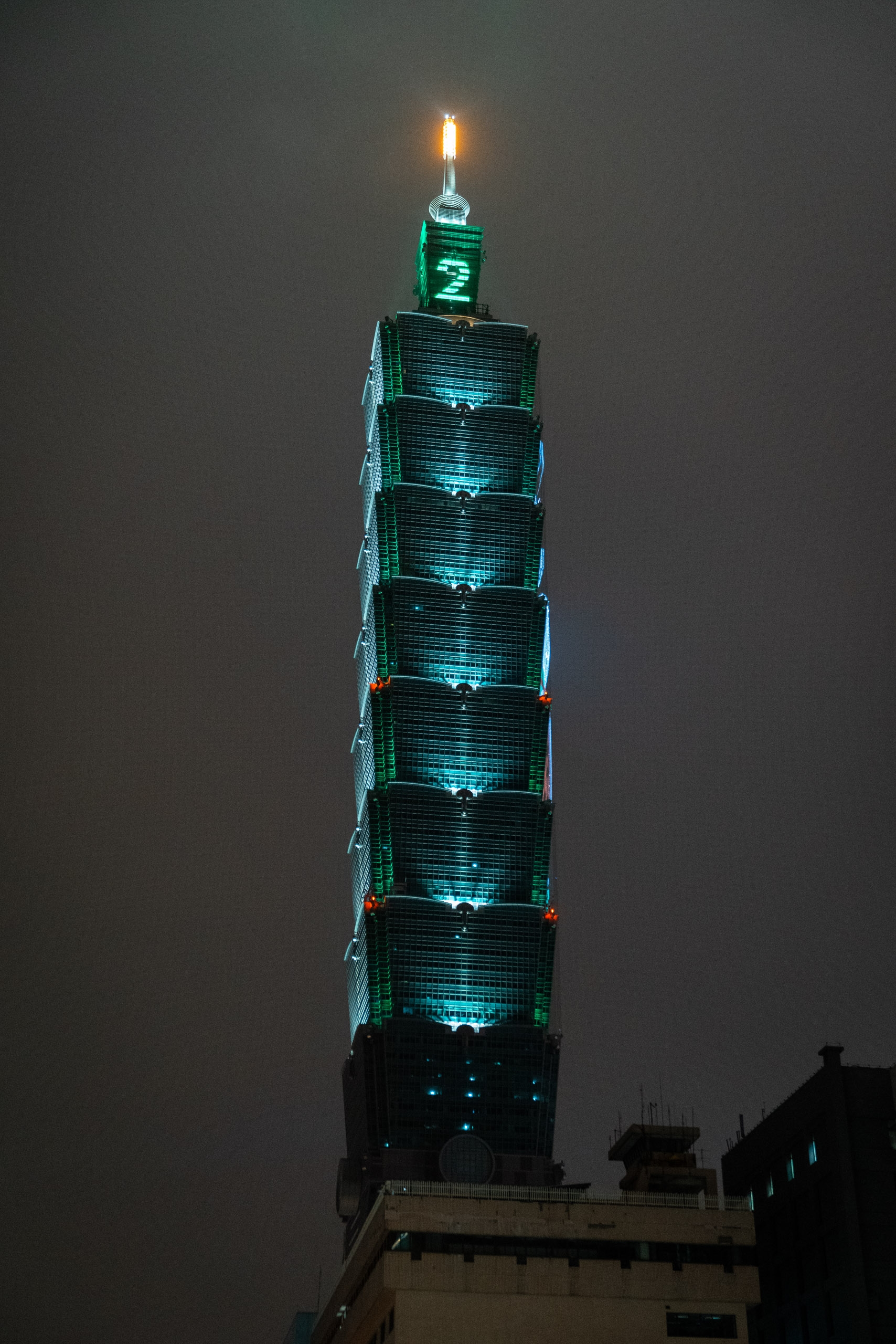 Taipei 101's illuminated spire rises against a dark gray night sky, its stacked cylindrical sections glowing with turquoise-blue lighting and a golden beacon at the top