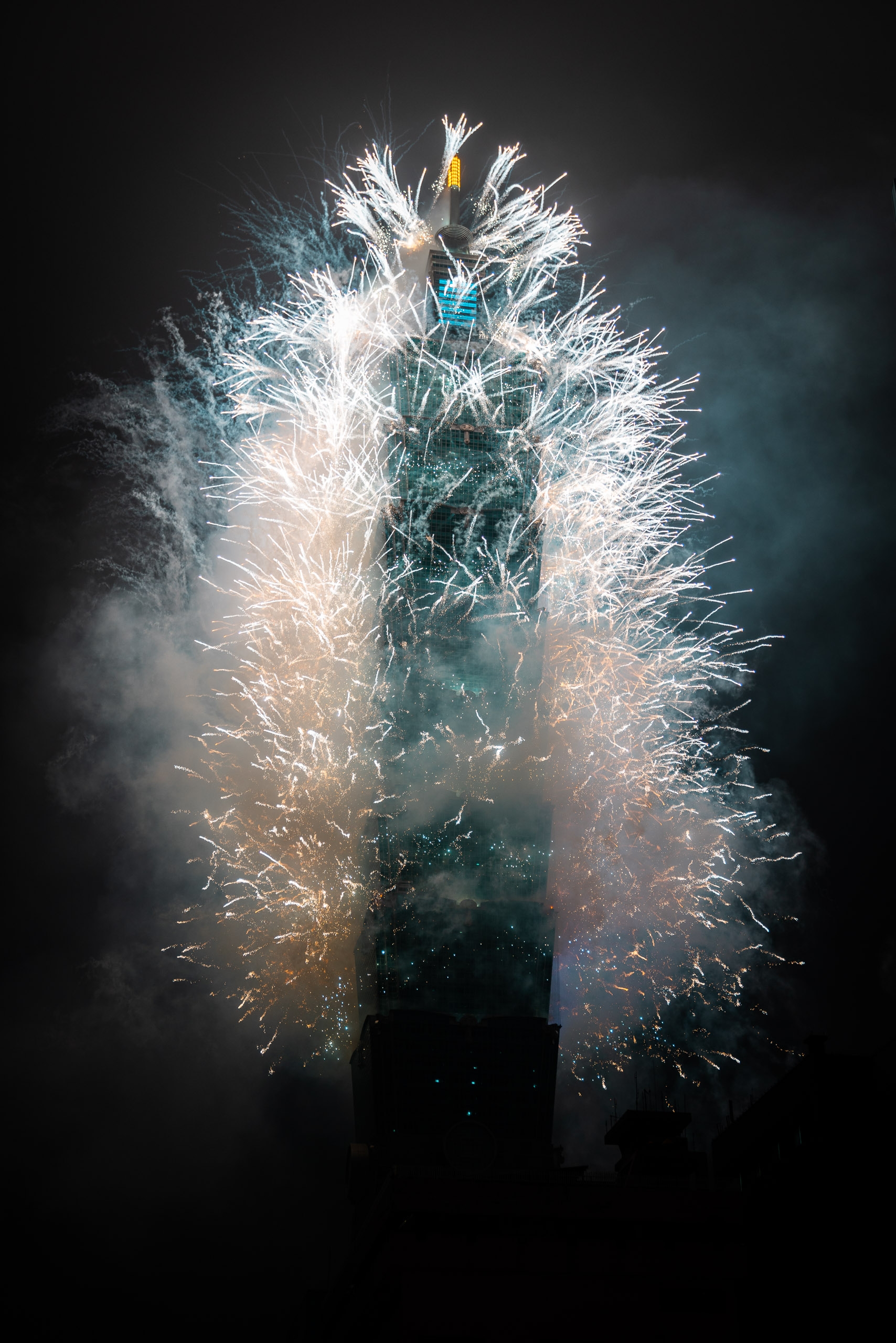 Taipei 101 tower illuminated with blue and cyan lights surrounded by exploding gold and purple fireworks against a dark night sky