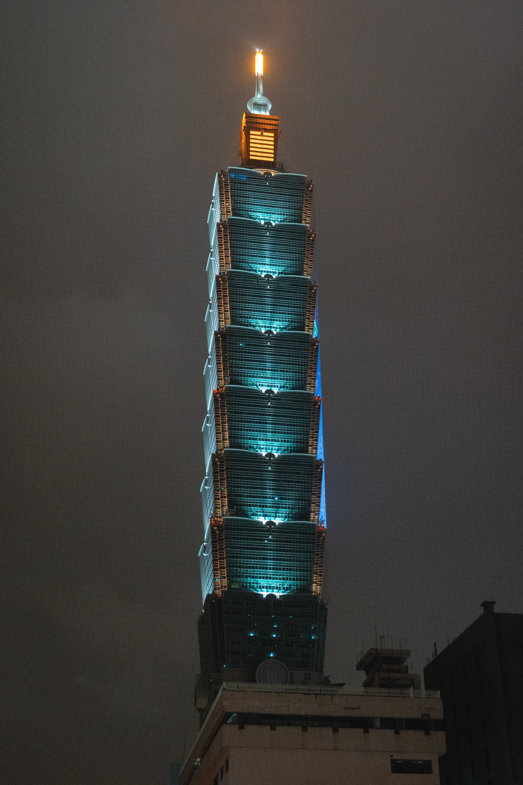 Taipei 101 tower illuminated at night with turquoise lighting along its segmented facade and a glowing golden spire extending into the dark gray sky