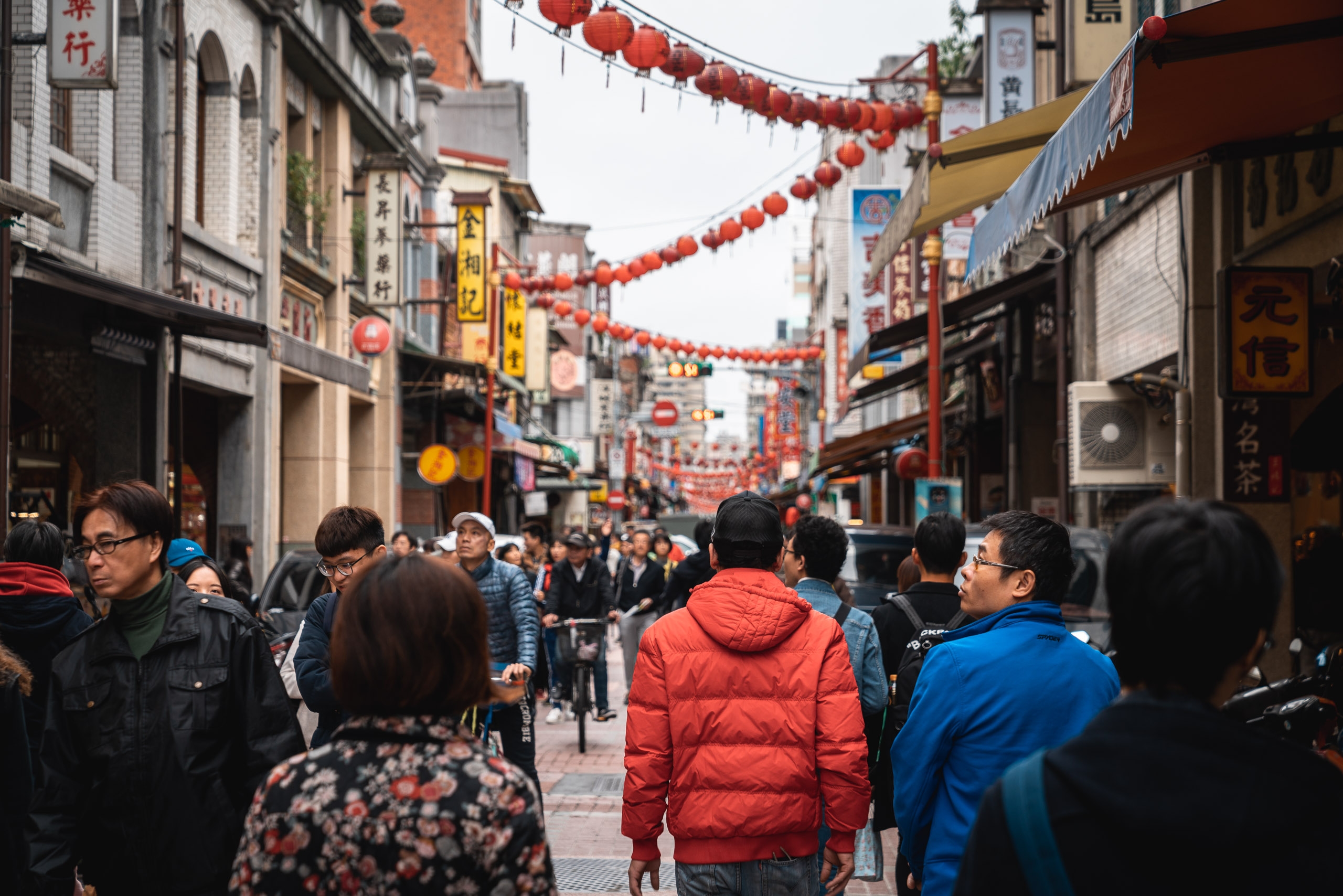 Crowded pedestrian street in a Taipei neighborhood decorated with hanging red lanterns, lined with traditional shopfronts and Chinese signage during daytime