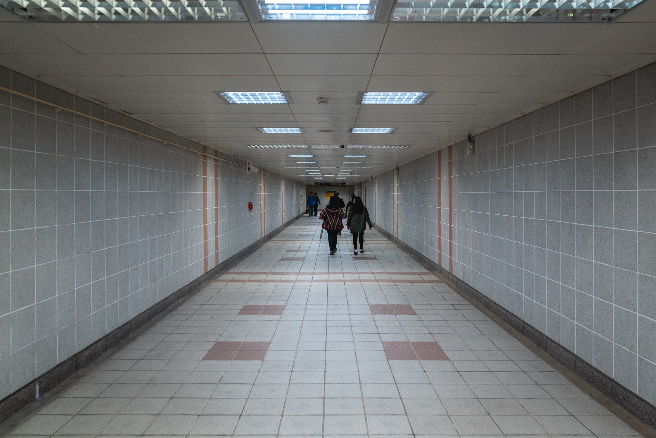 A tiled underground subway tunnel stretches into the distance with fluorescent ceiling lights and a few pedestrians walking through the empty passage