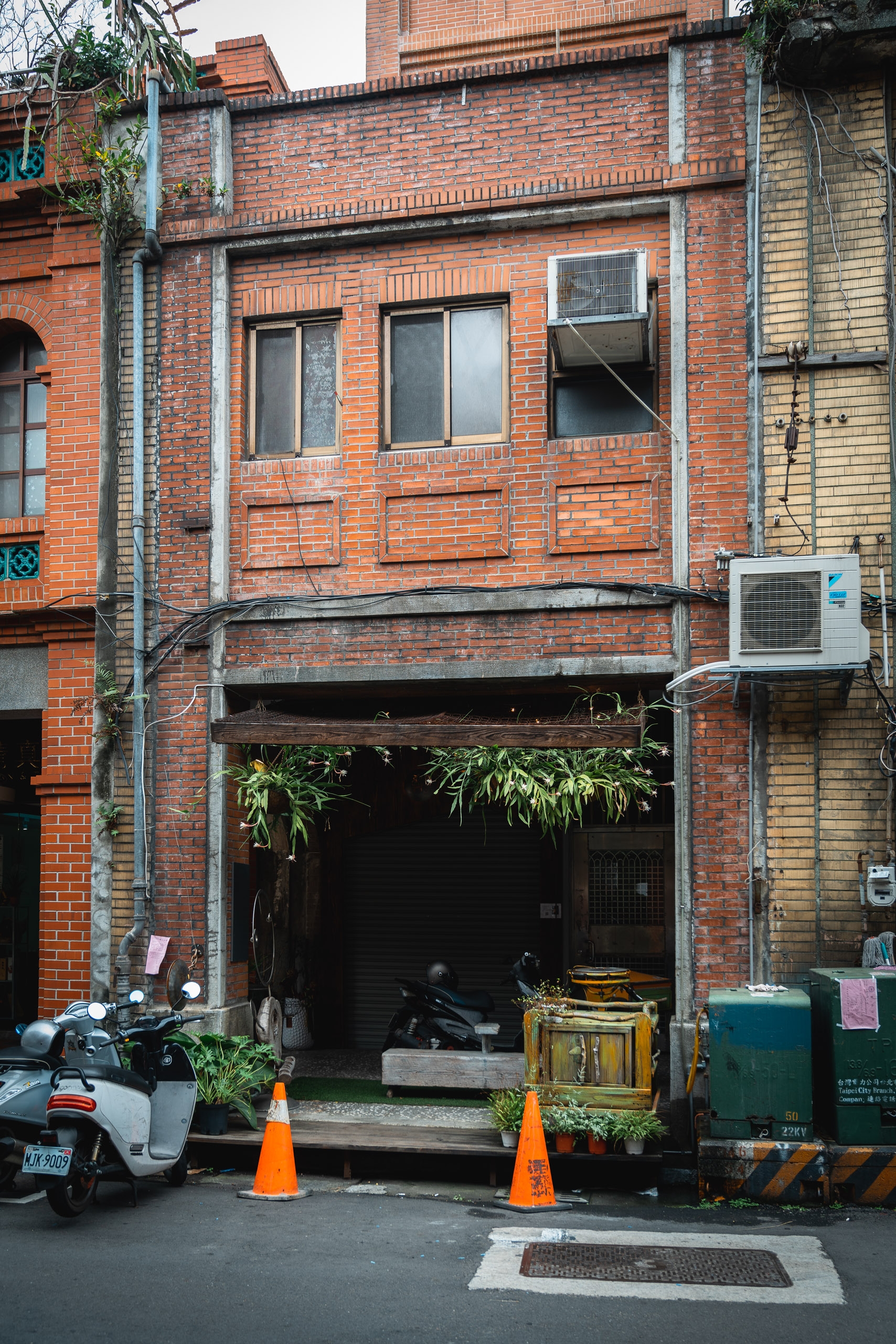 A weathered red-brick building with dark storefront windows sits on a narrow Taipei street, flanked by orange traffic cones and a parked scooter, with utility units and climbing vines visible on the facade