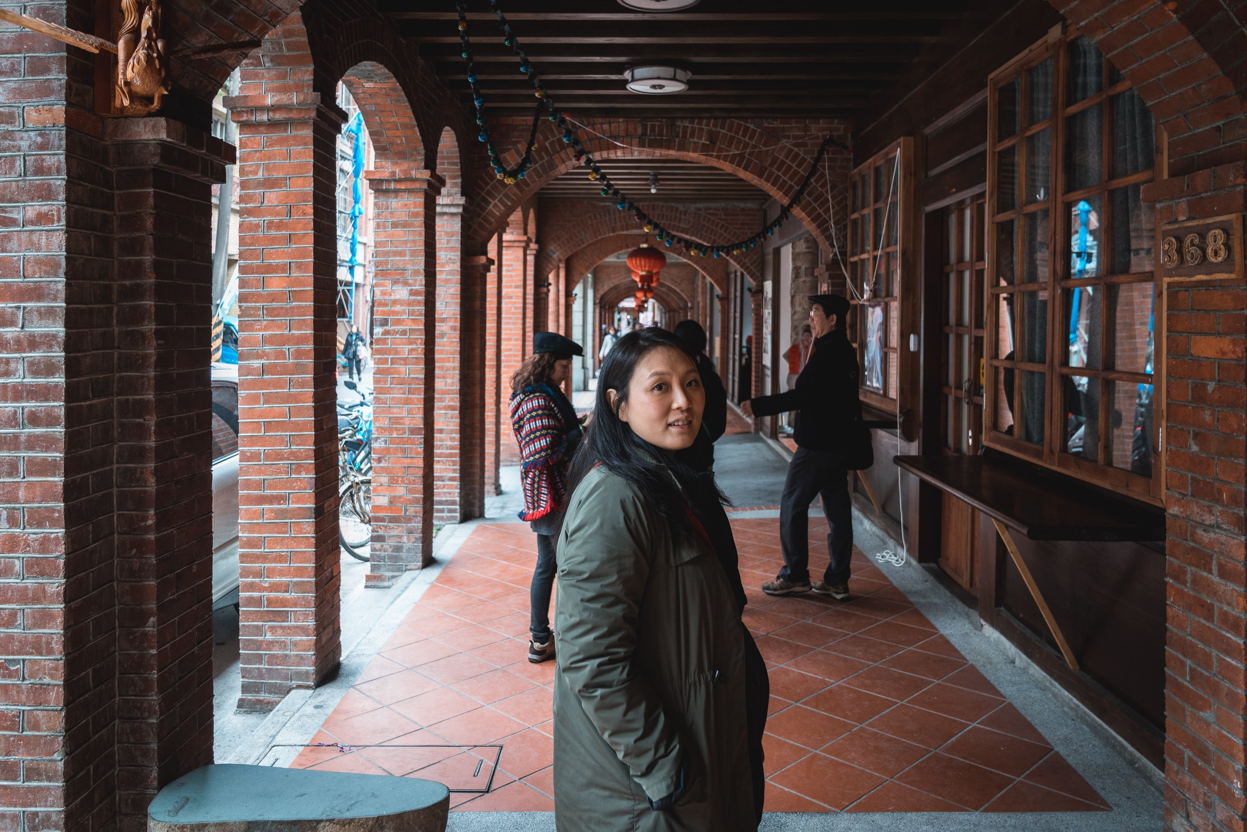 A woman in a green jacket walks through a narrow brick-arched corridor lined with wooden storefronts and red tile flooring, with other visitors visible in the traditional Taiwanese alleyway