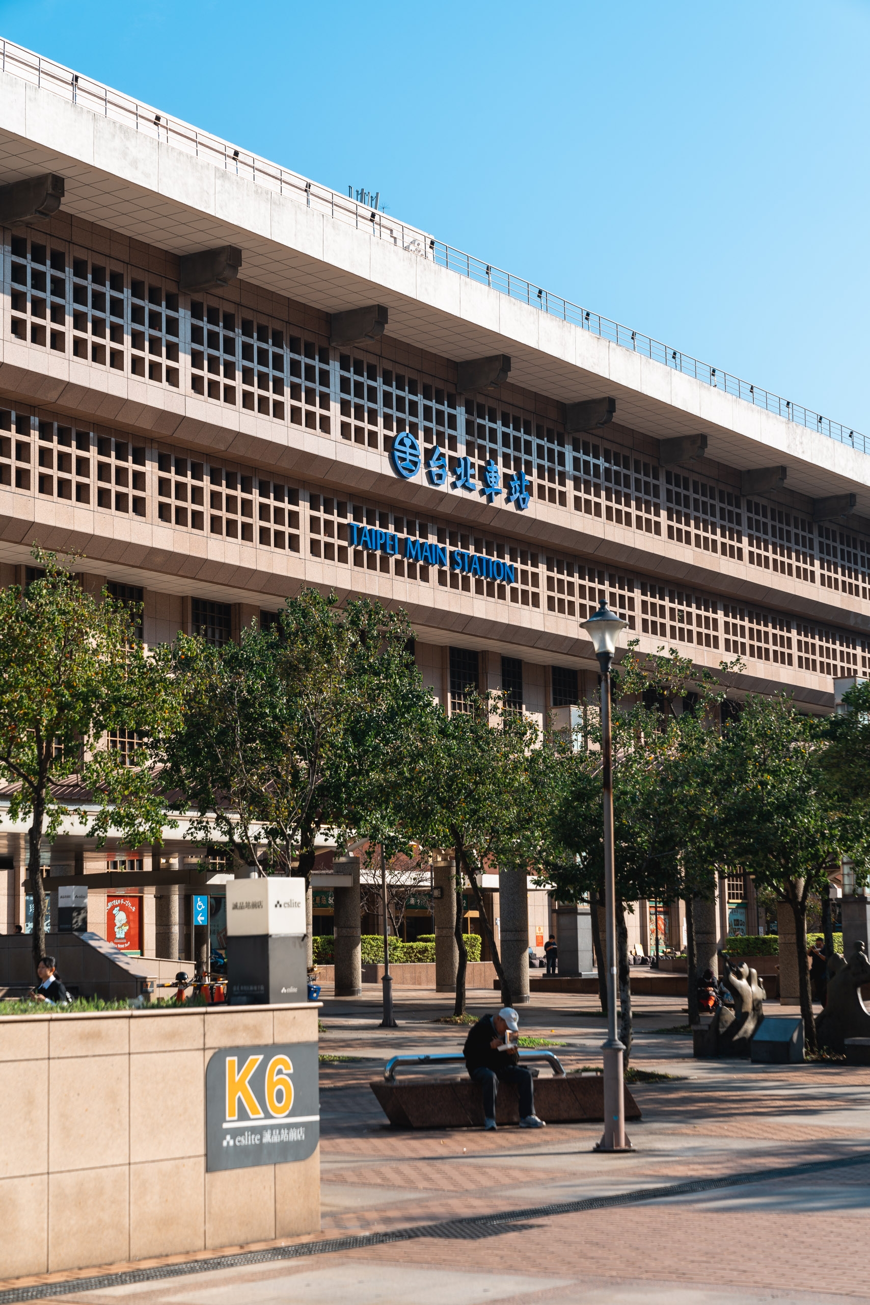 A brutalist concrete building with blue signage rises above a sun-drenched plaza featuring green trees, street-level shops, and a K6 directional marker under clear blue skies