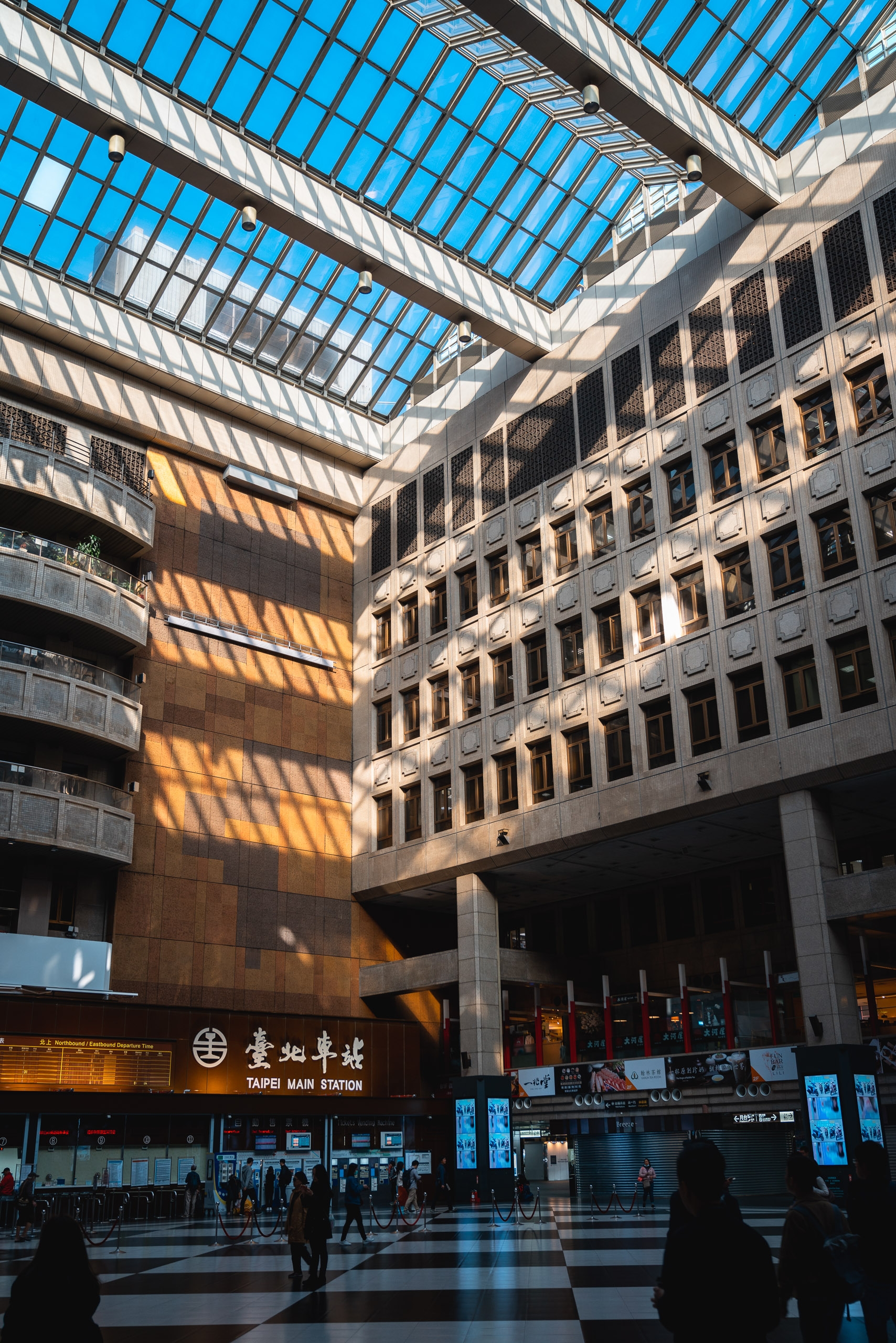 Taipei Main Station's modern courtyard features glass skylight panels casting geometric shadows across rust-toned brick walls and the bustling plaza below on a clear day