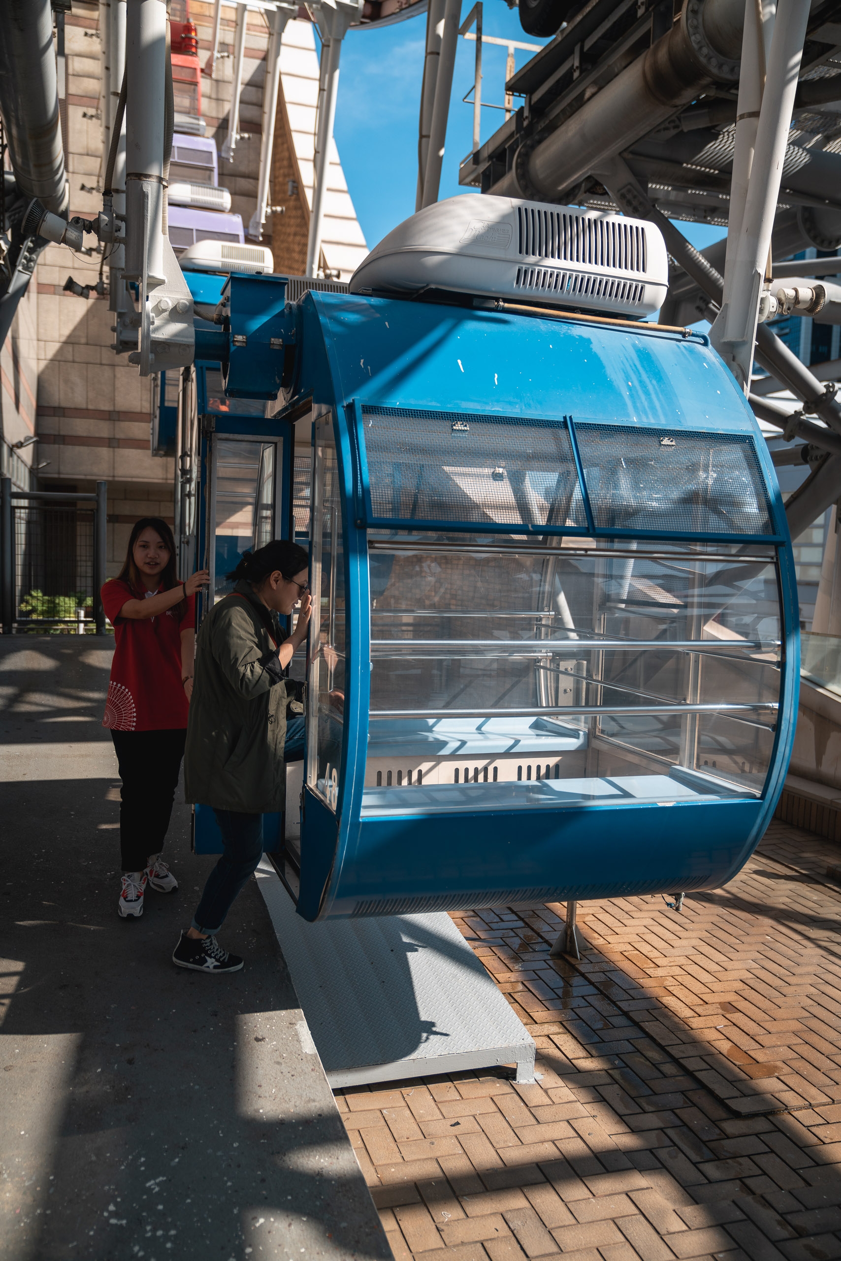 Two passengers stand at the entrance of a bright blue cable car cabin with clear windows, positioned in an urban setting with brick pavement and metal support structures overhead