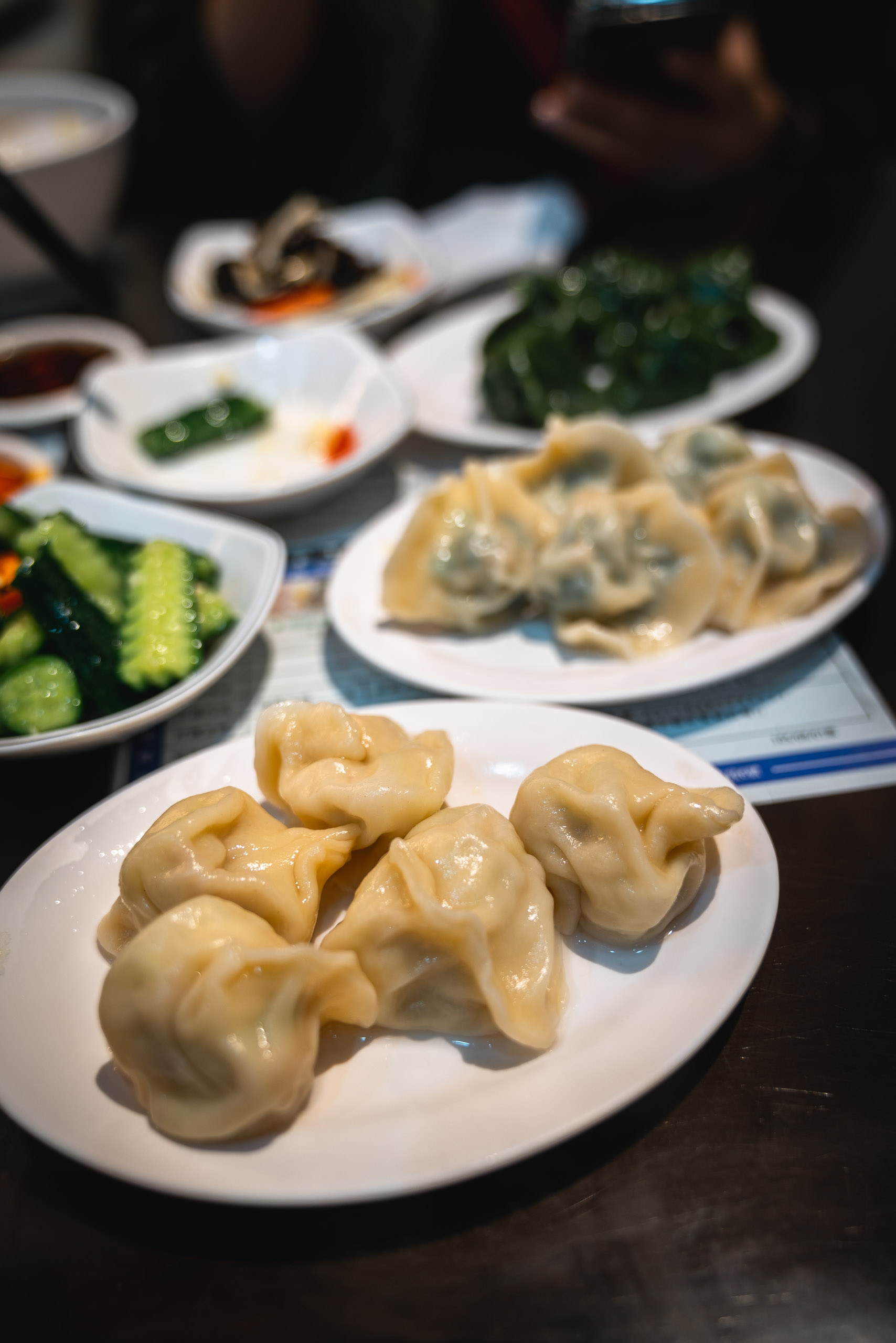 Steamed dumplings and vegetable dishes arranged on white plates at a dimly lit Asian restaurant table with diners in the background