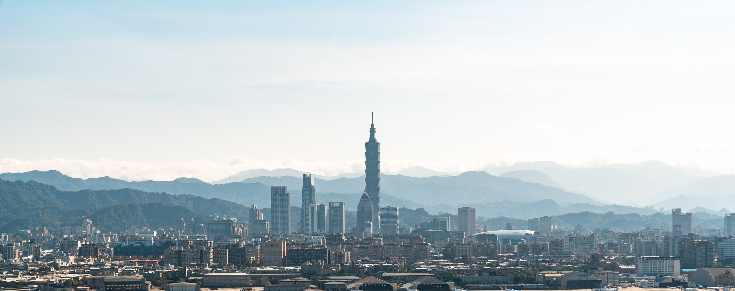 Taipei's skyline rises dramatically against distant mountains under hazy daylight, with Taipei 101's distinctive spire dominating the center of the dense urban landscape