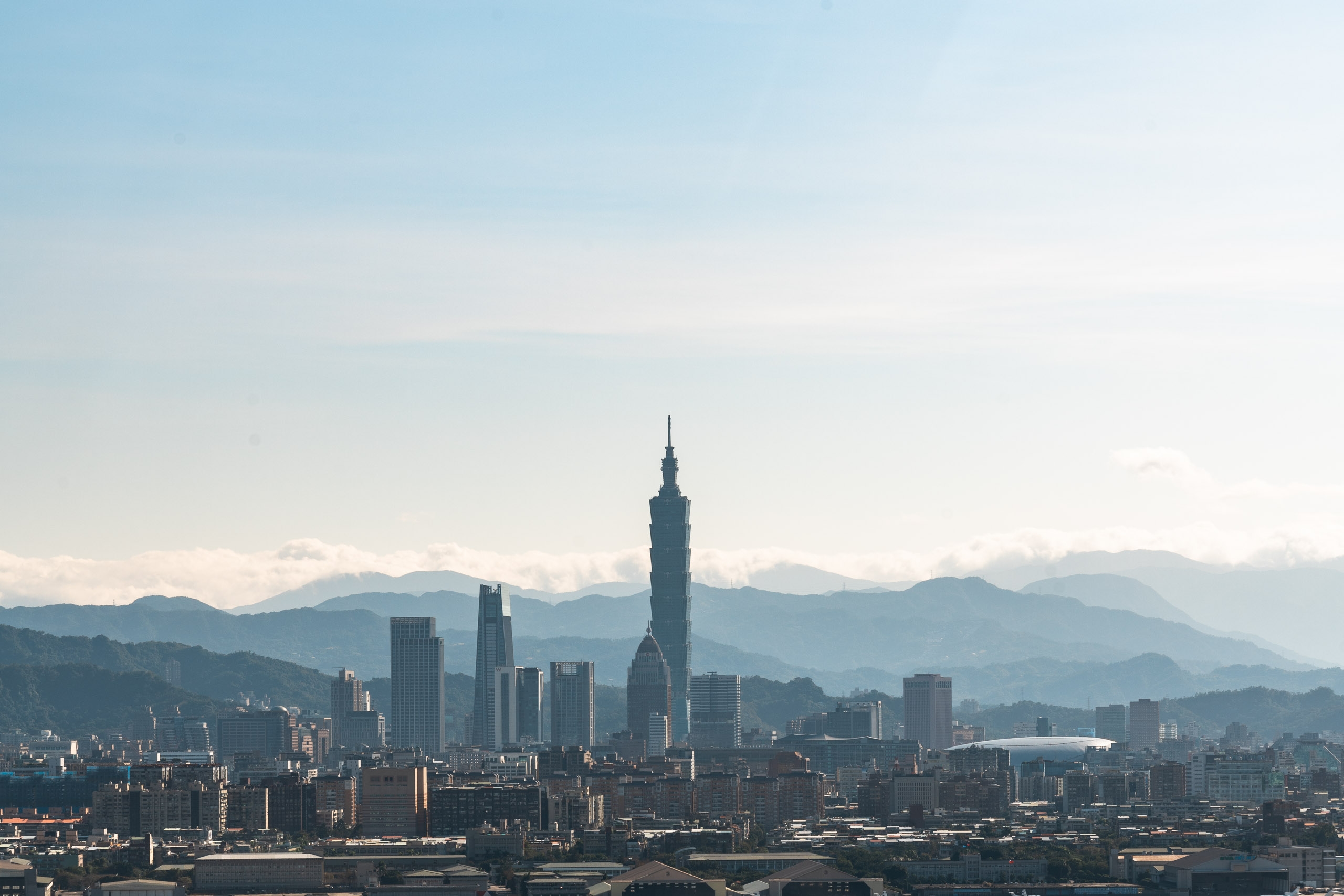 Taipei's skyline features Taipei 101 tower rising prominently above surrounding buildings with misty mountains visible in the hazy background under clear daylight