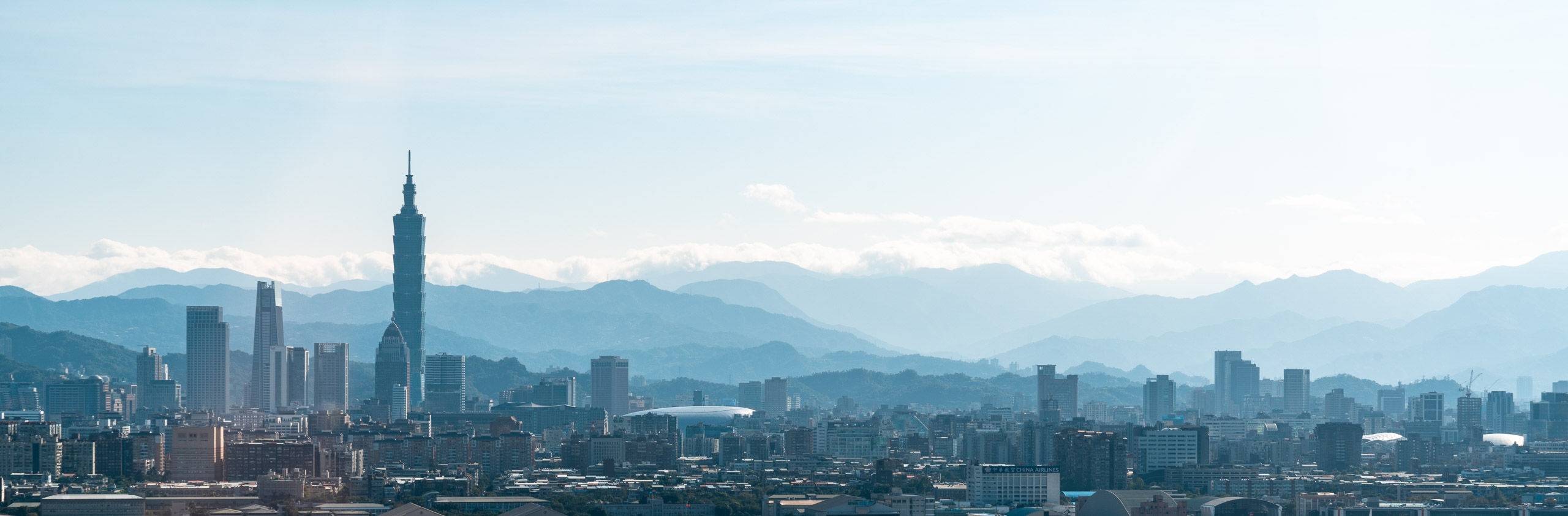 Taipei's skyline stretches across the frame with Taipei 101 rising prominently above surrounding buildings, mountains visible hazy in the distance under overcast skies