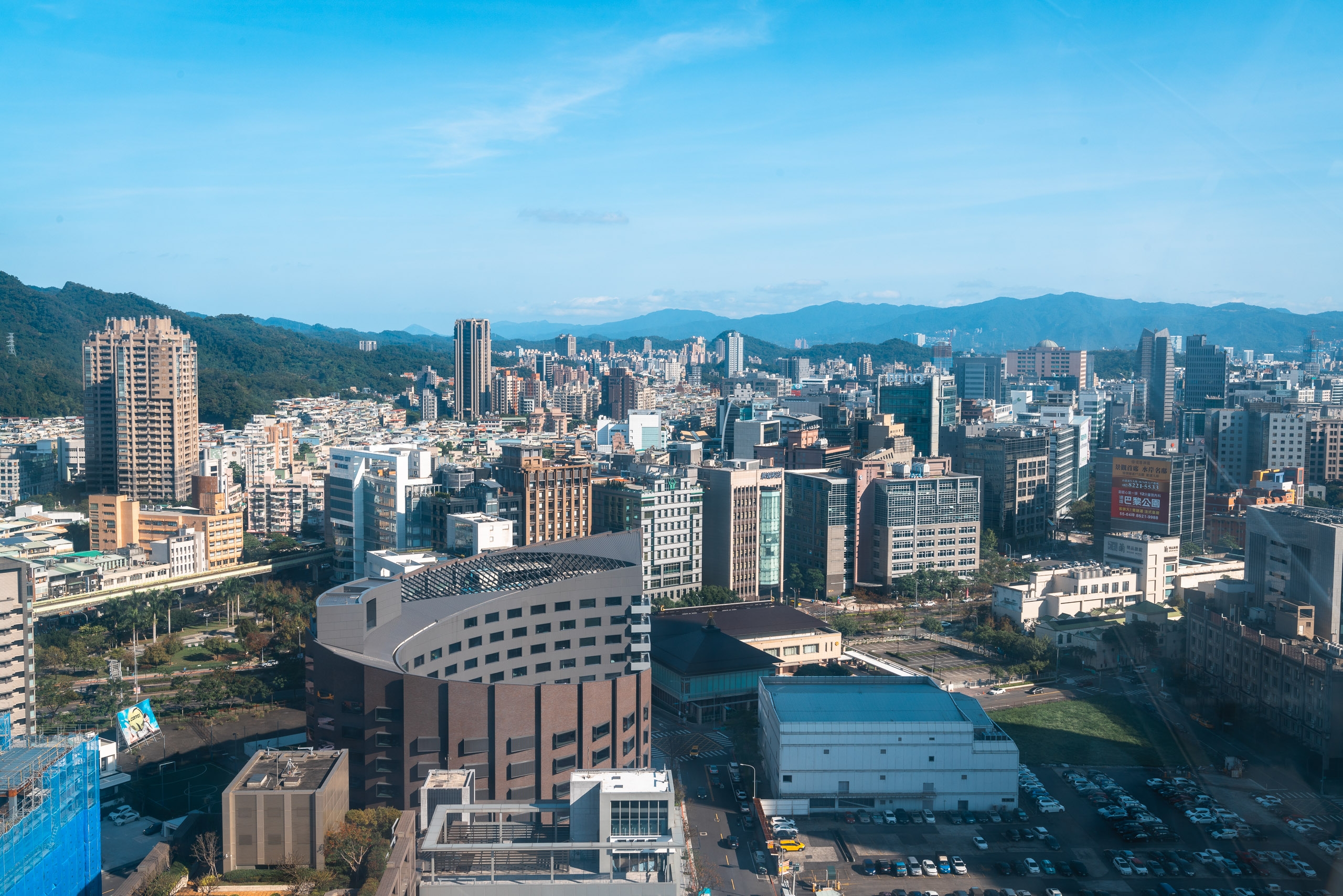 Aerial view of Taipei's dense urban skyline with modern high-rises, a distinctive curved building in the foreground, mountains rising beyond the city, and clear blue sky overhead