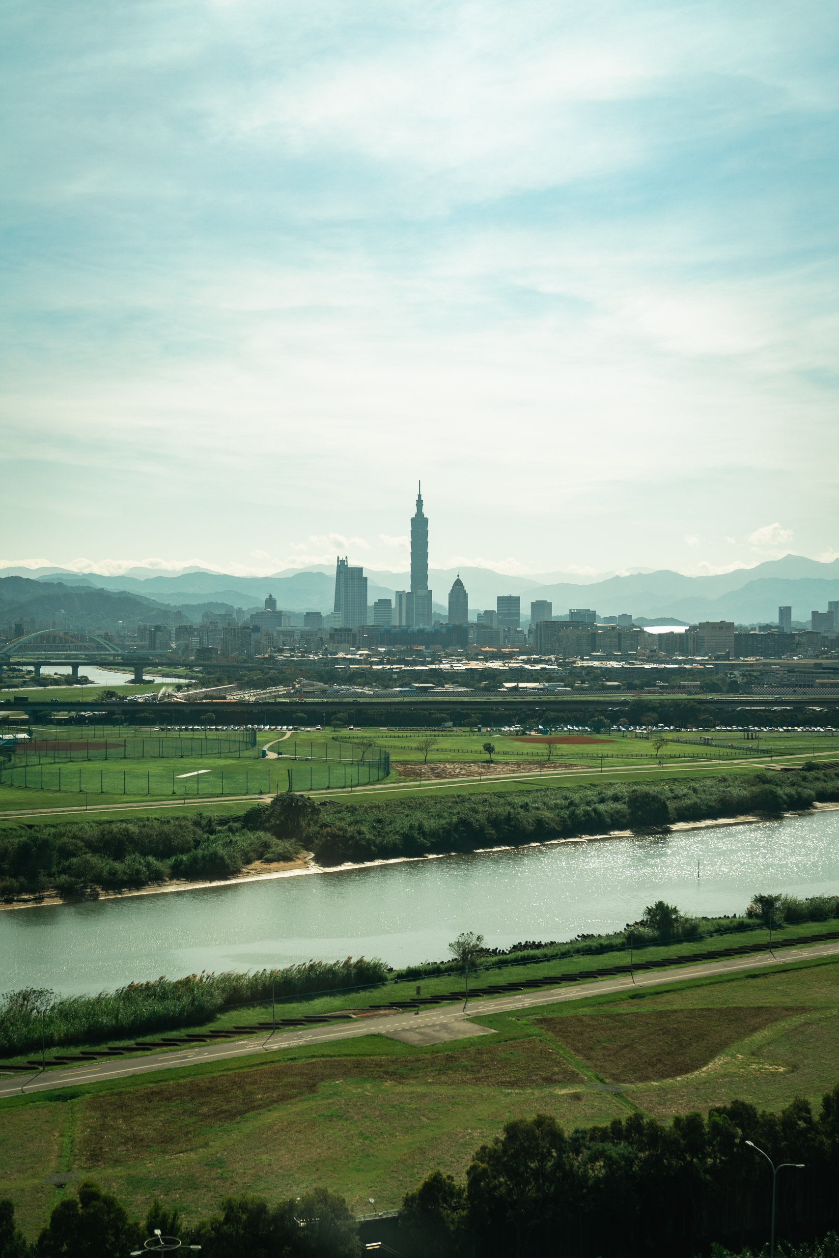 Taipei's skyline rises behind agricultural fields and a winding river under hazy daylight, with distant mountains framing the urban landscape