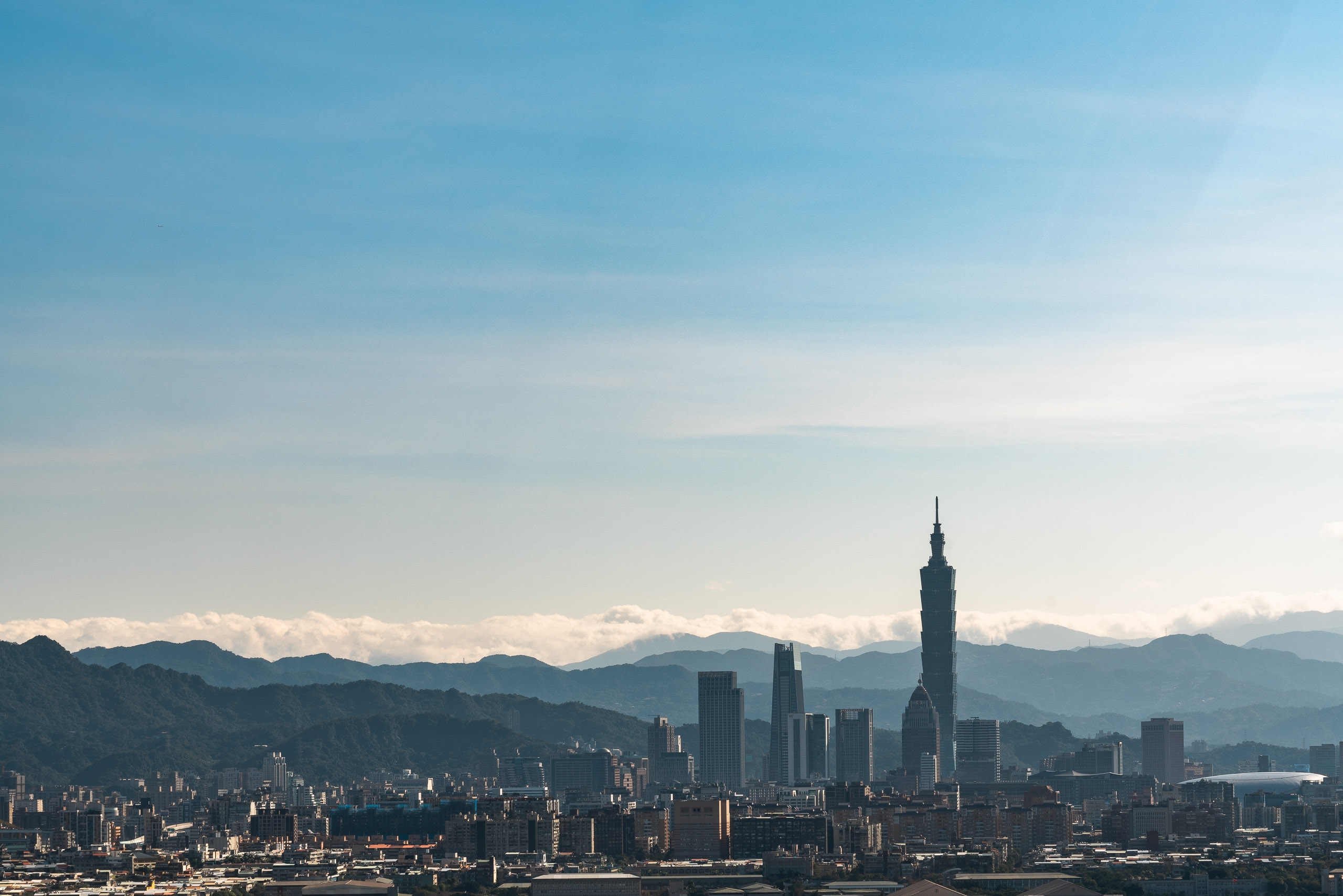Taipei's skyline stretches across the frame with Taipei 101 dominating the center, mountains rising in the hazy distance under clear blue sky