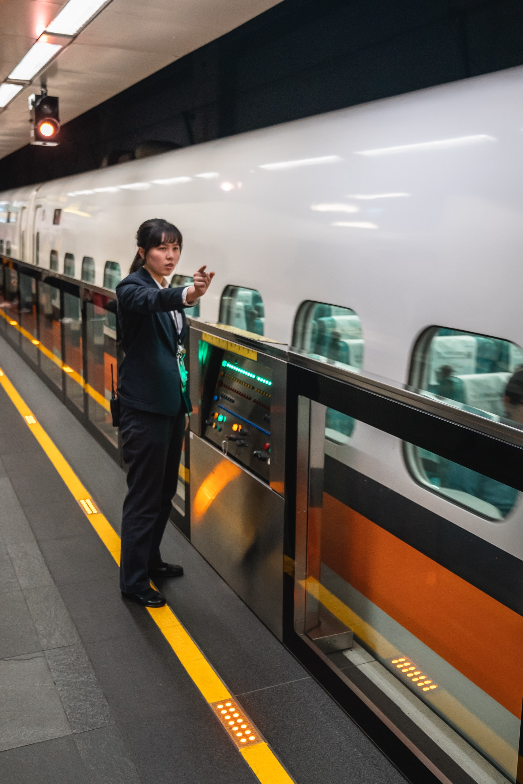 A transit worker in dark uniform stands beside an orange and white train at a modern Taipei subway platform, with fluorescent lighting overhead and yellow safety markings on the floor