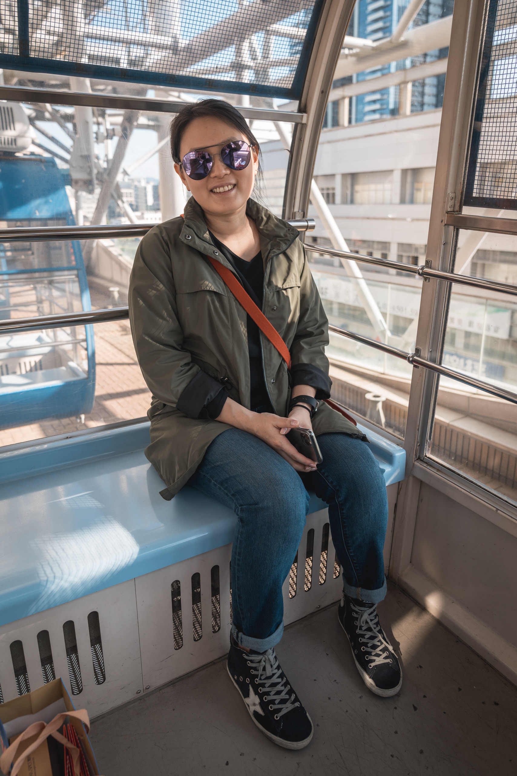 A woman wearing sunglasses and a olive jacket sits on a blue bench inside a cable car cabin, with urban buildings visible through the windows behind her