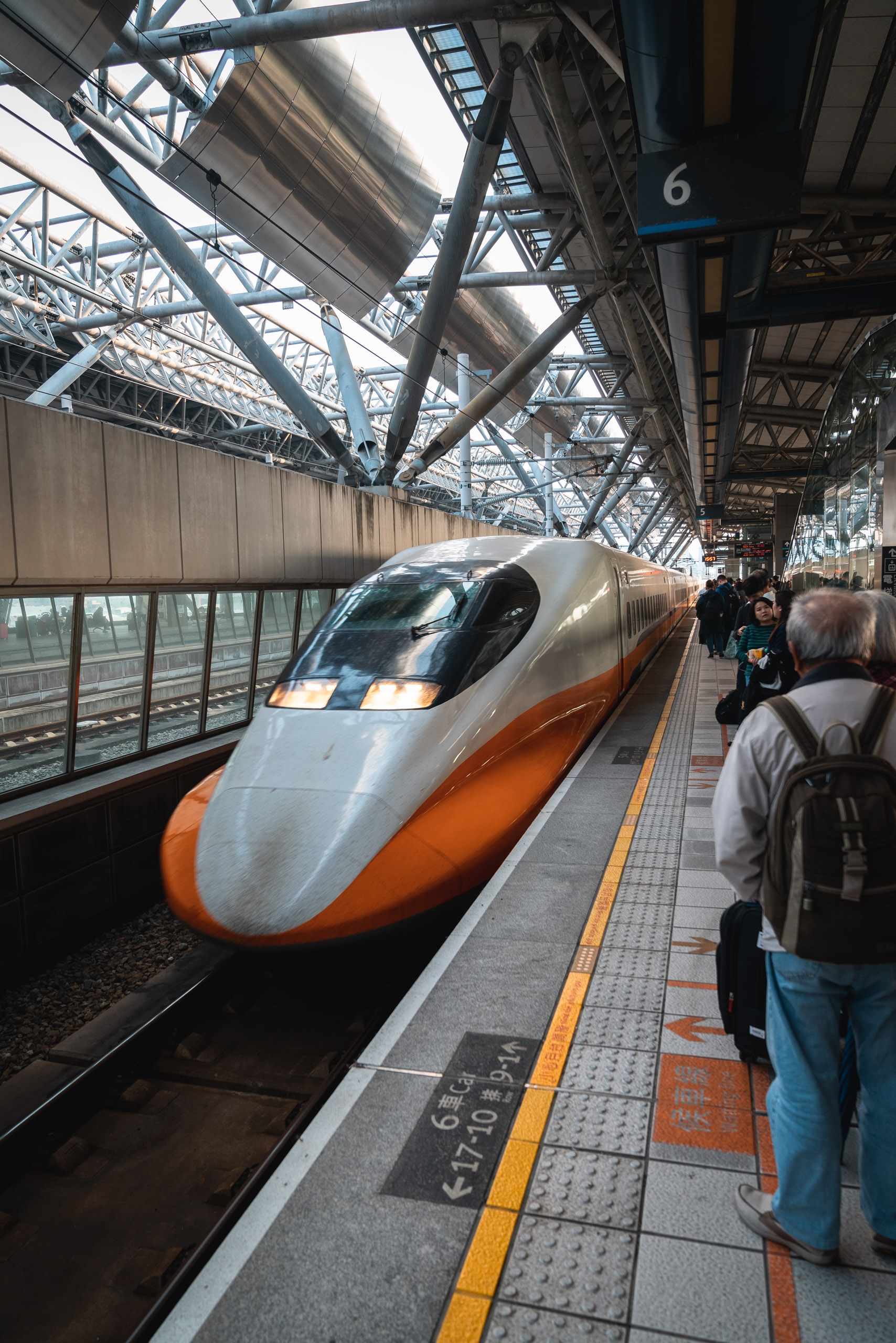 A sleek orange-and-white high-speed train sits at platform six beneath an industrial steel framework, with passengers waiting alongside the tactile-patterned platform edge