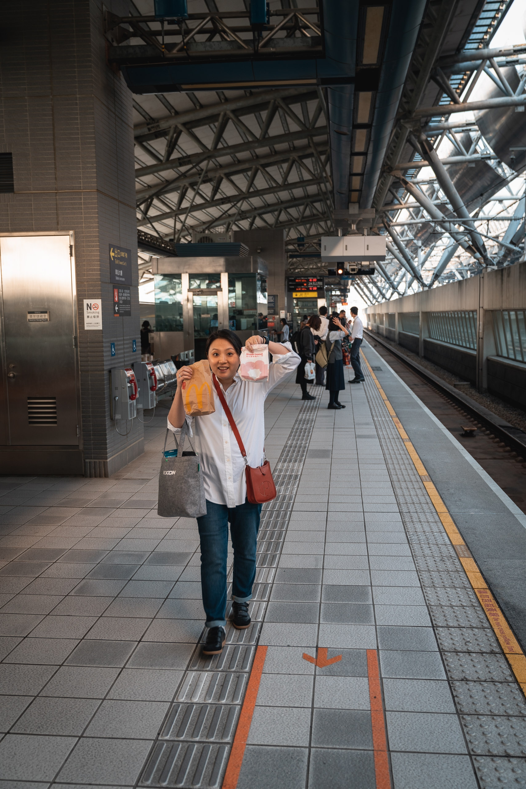 A woman in a white shirt holds up a pink shopping bag while smiling at the camera on a modern Japanese train platform with metal roof structures and other passengers waiting in the background.