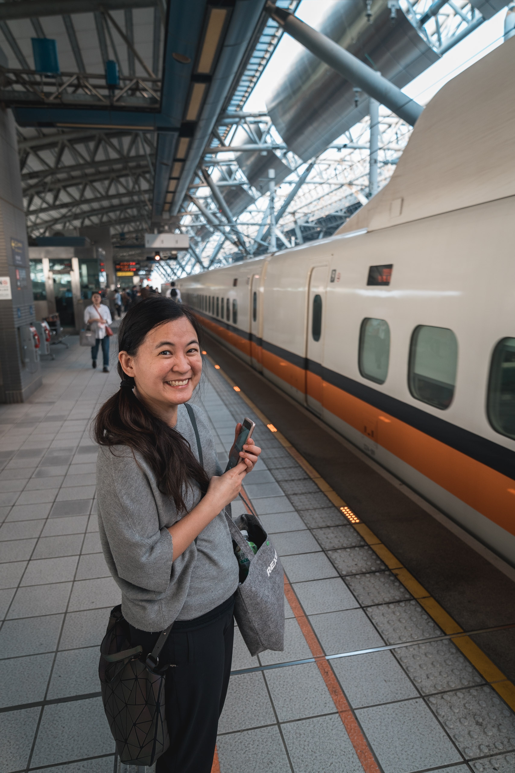 A smiling woman stands on a train platform beside a modern white and orange high-speed train, holding a ticket while looking back at the camera under the station's industrial metal roof structure