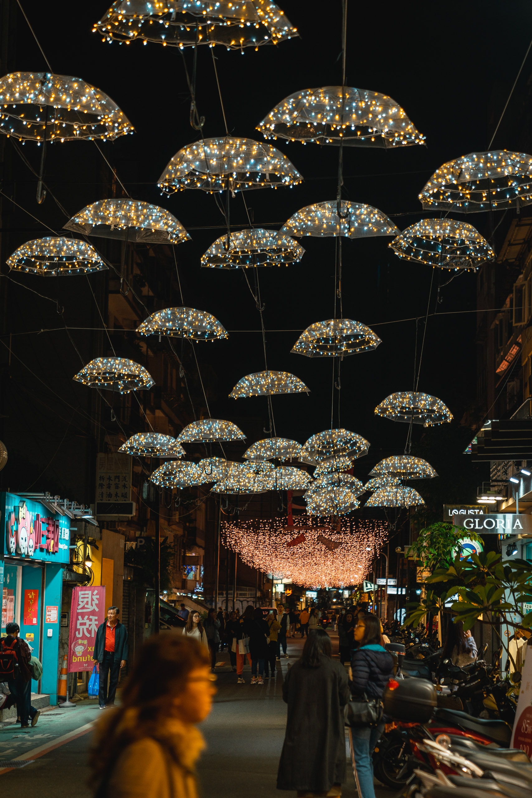 Illuminated umbrellas strung overhead create a canopy of warm lights above a bustling night market street in Taipei, with neon signs and pedestrians visible below