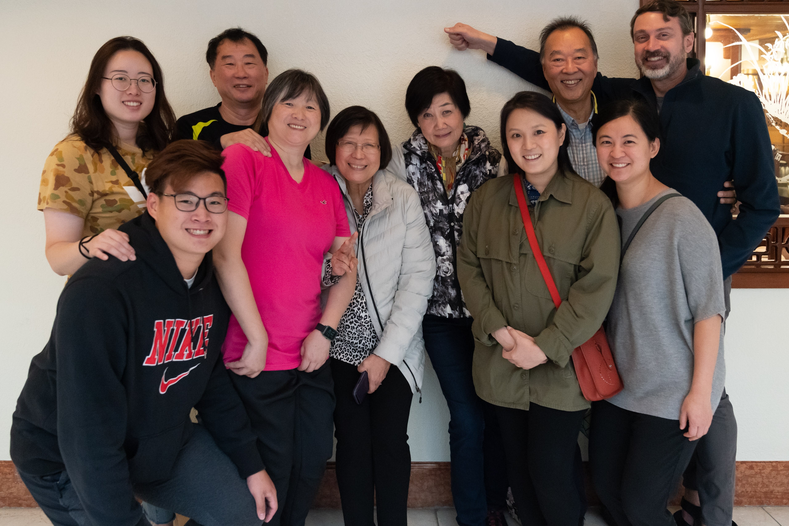 A diverse group of nine people of varying ages stands together against a white wall, smiling warmly at the camera in an indoor setting with warm lighting from a window visible behind them.