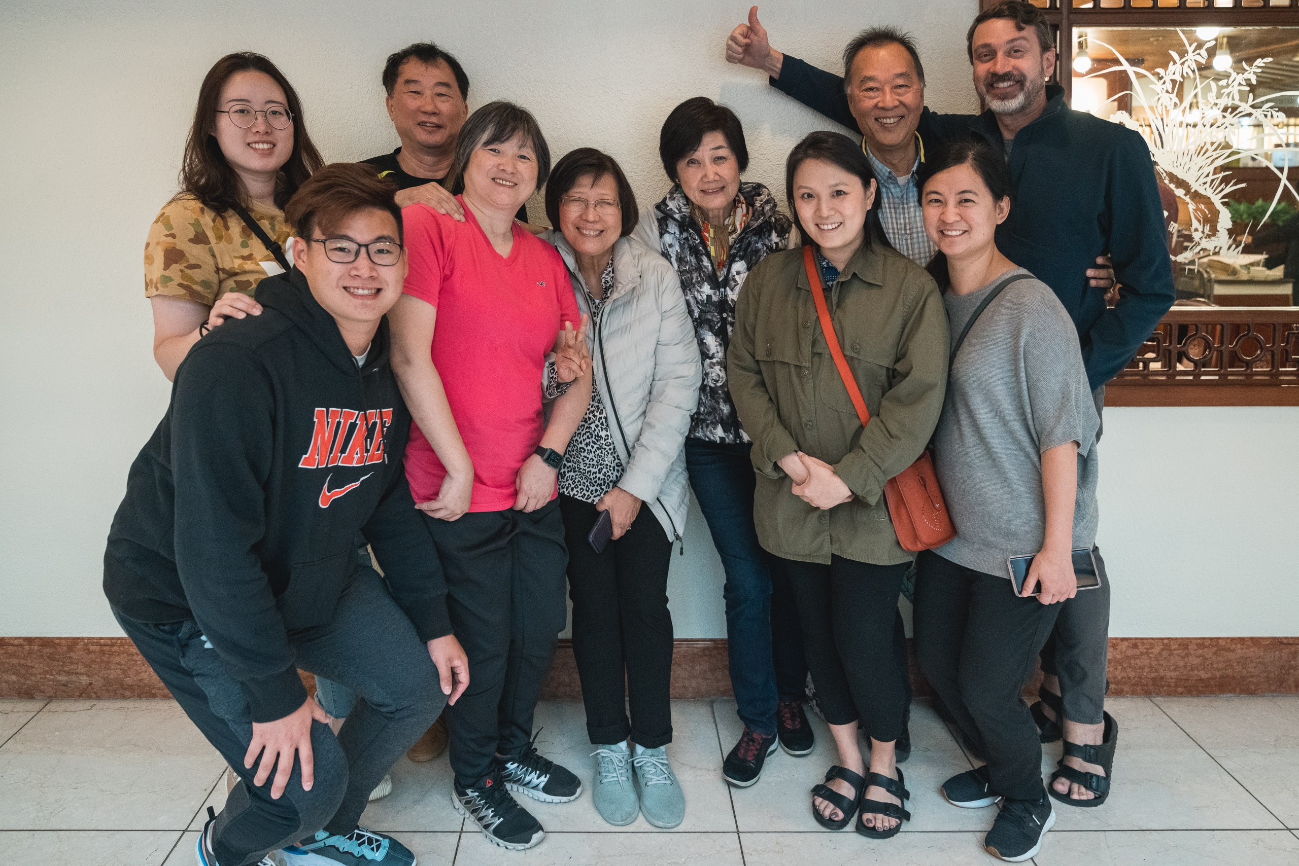 A multi-generational family group of nine people poses together indoors against a white wall, smiling at the camera in casual clothing