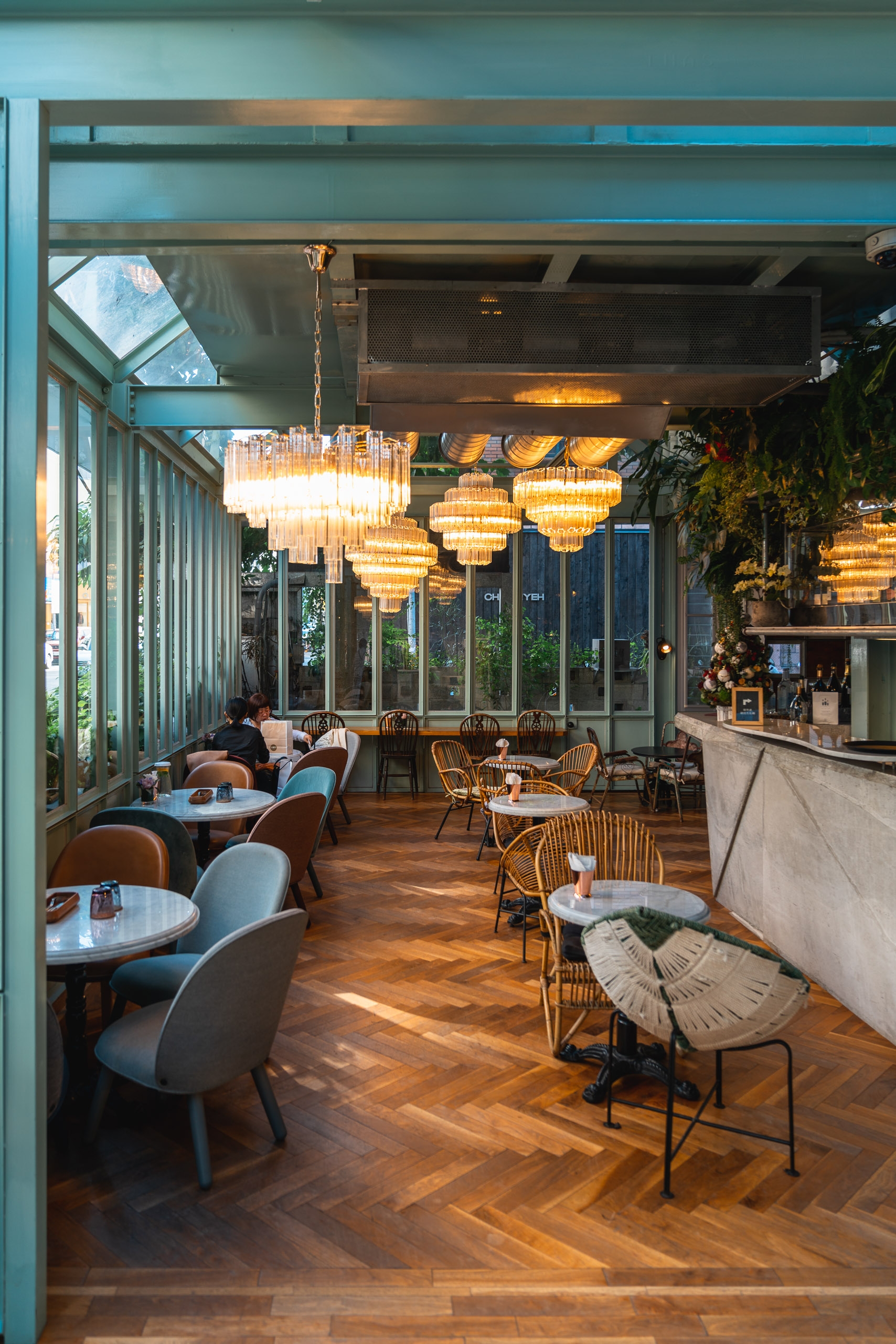 Modern restaurant interior with wooden herringbone flooring, tiered brass pendant lights, and mid-century seating arranged throughout the bright, plant-adorned space with floor-to-ceiling windows