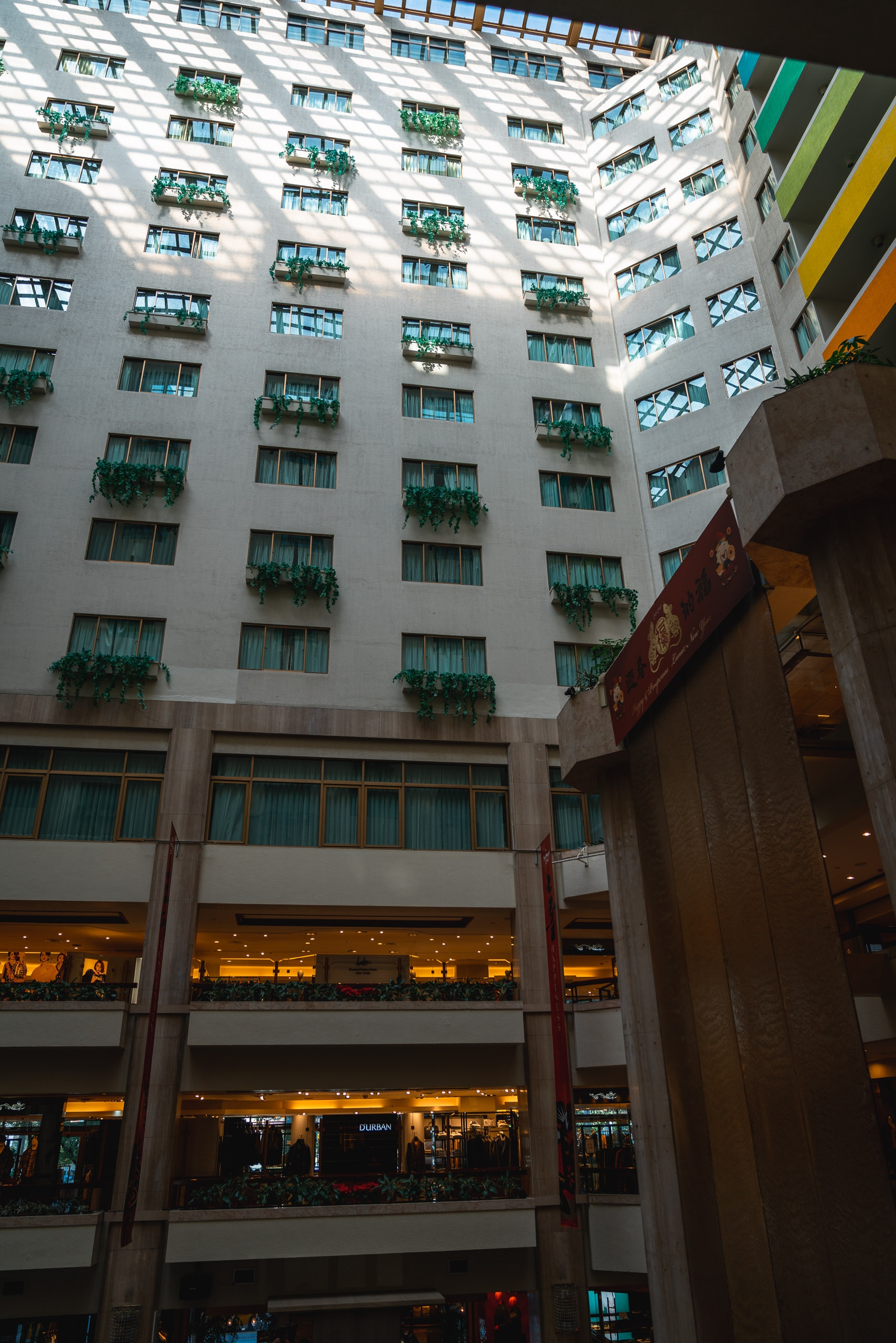 A white residential building with cascading green planters on each balcony rises above an illuminated shopping arcade, photographed from within the courtyard looking upward toward bright daylight