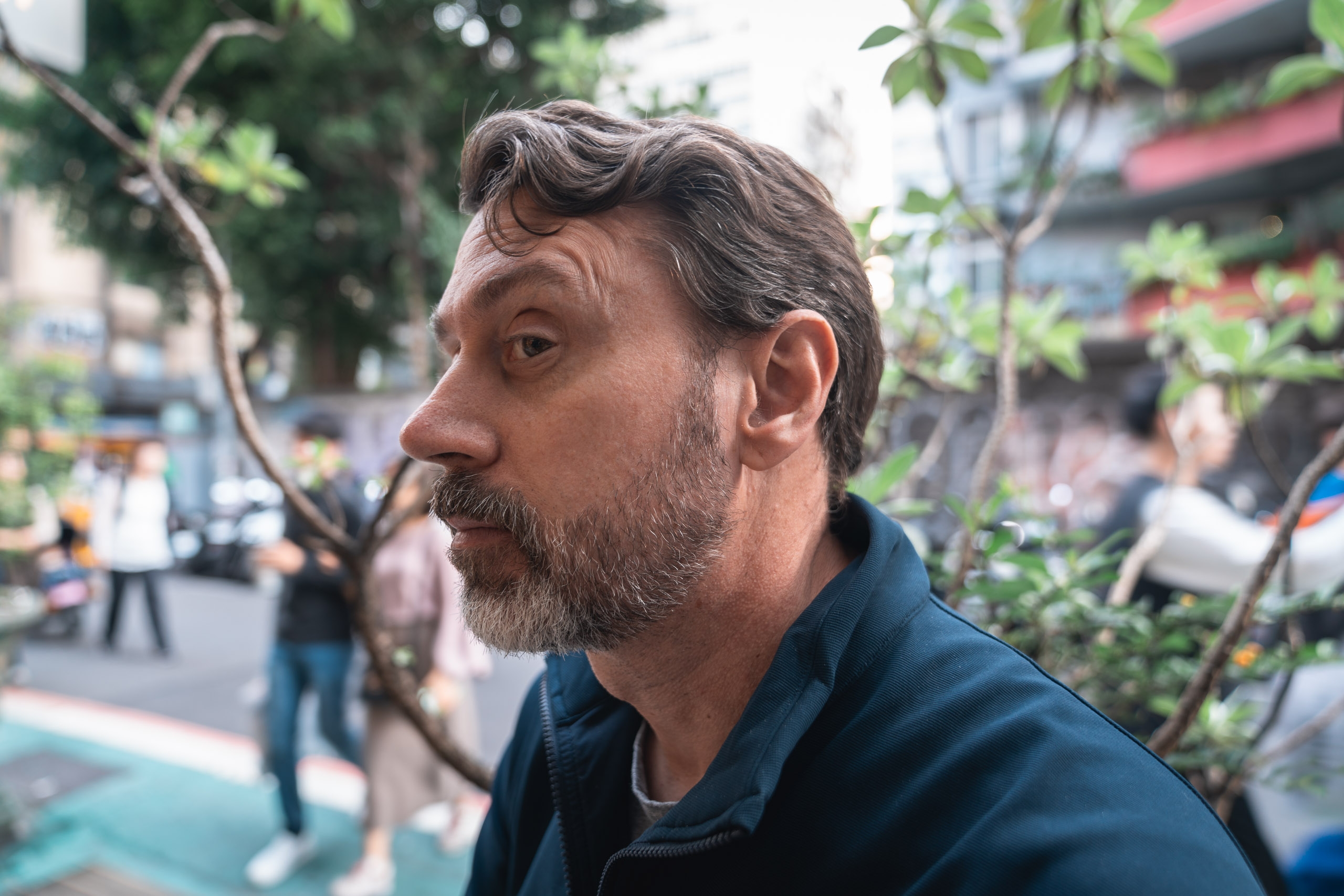 Bearded man in dark blue jacket gazes thoughtfully to the side on a busy Taipei street surrounded by trees and blurred pedestrians