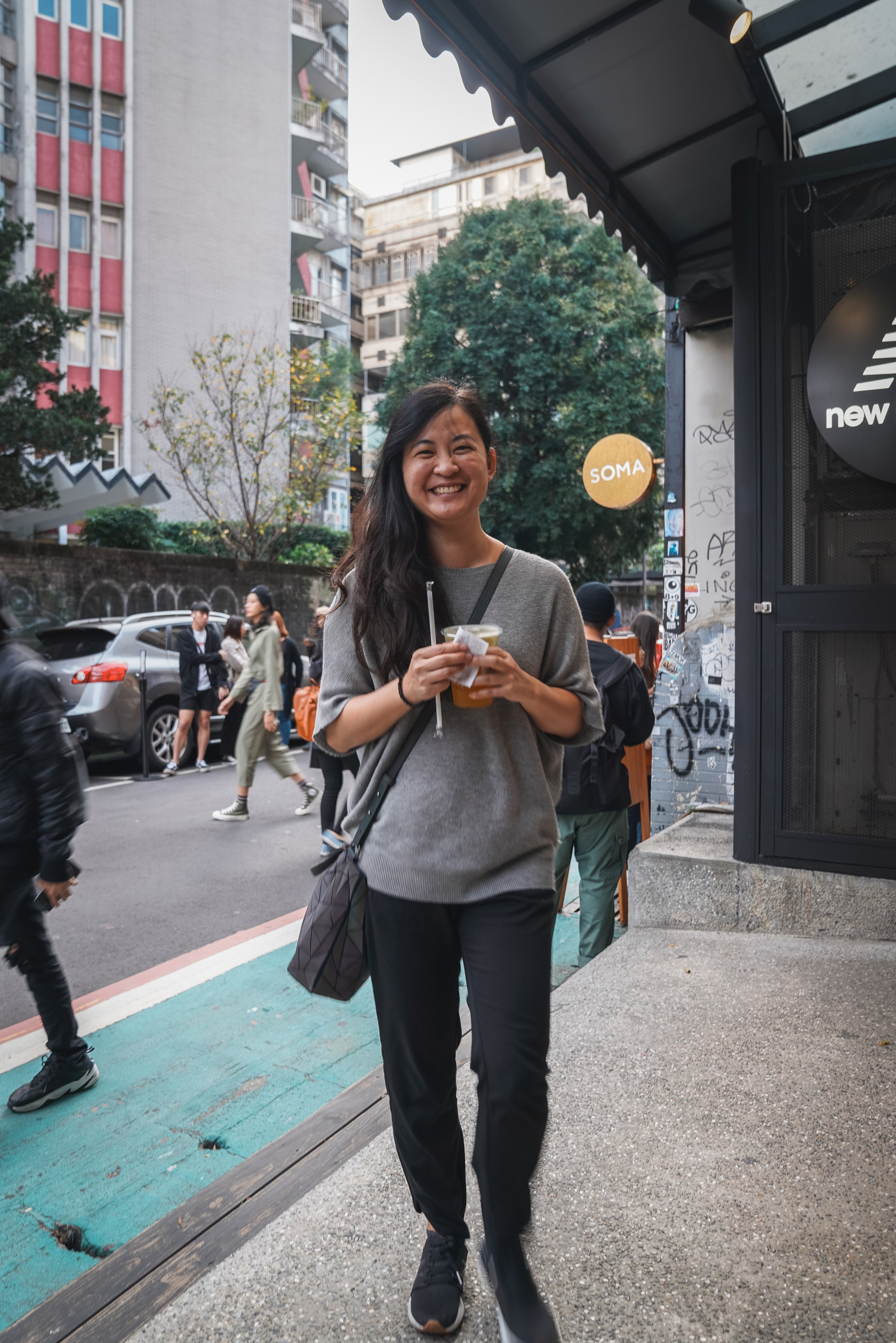 A smiling woman in a gray shirt holds a beverage while standing on a vibrant turquoise sidewalk outside a modern storefront, surrounded by urban Taipei architecture and pedestrians