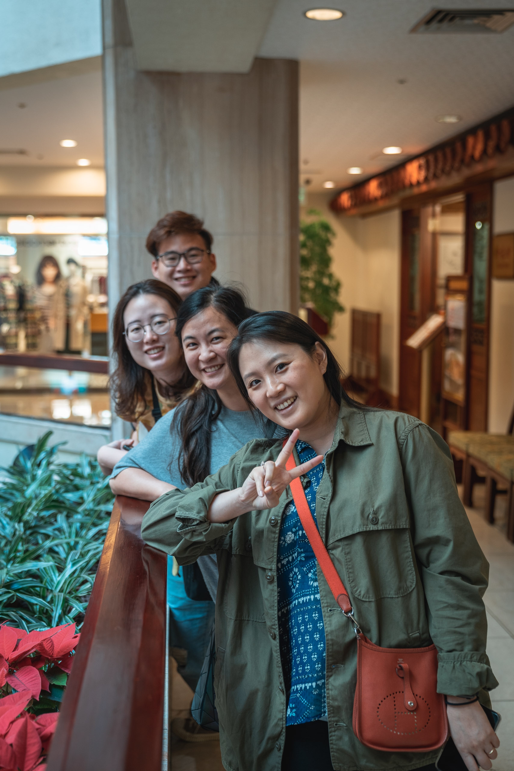Four young people pose together in a modern shopping mall corridor, smiling and making peace signs, with potted plants and warm overhead lighting framing the indoor scene