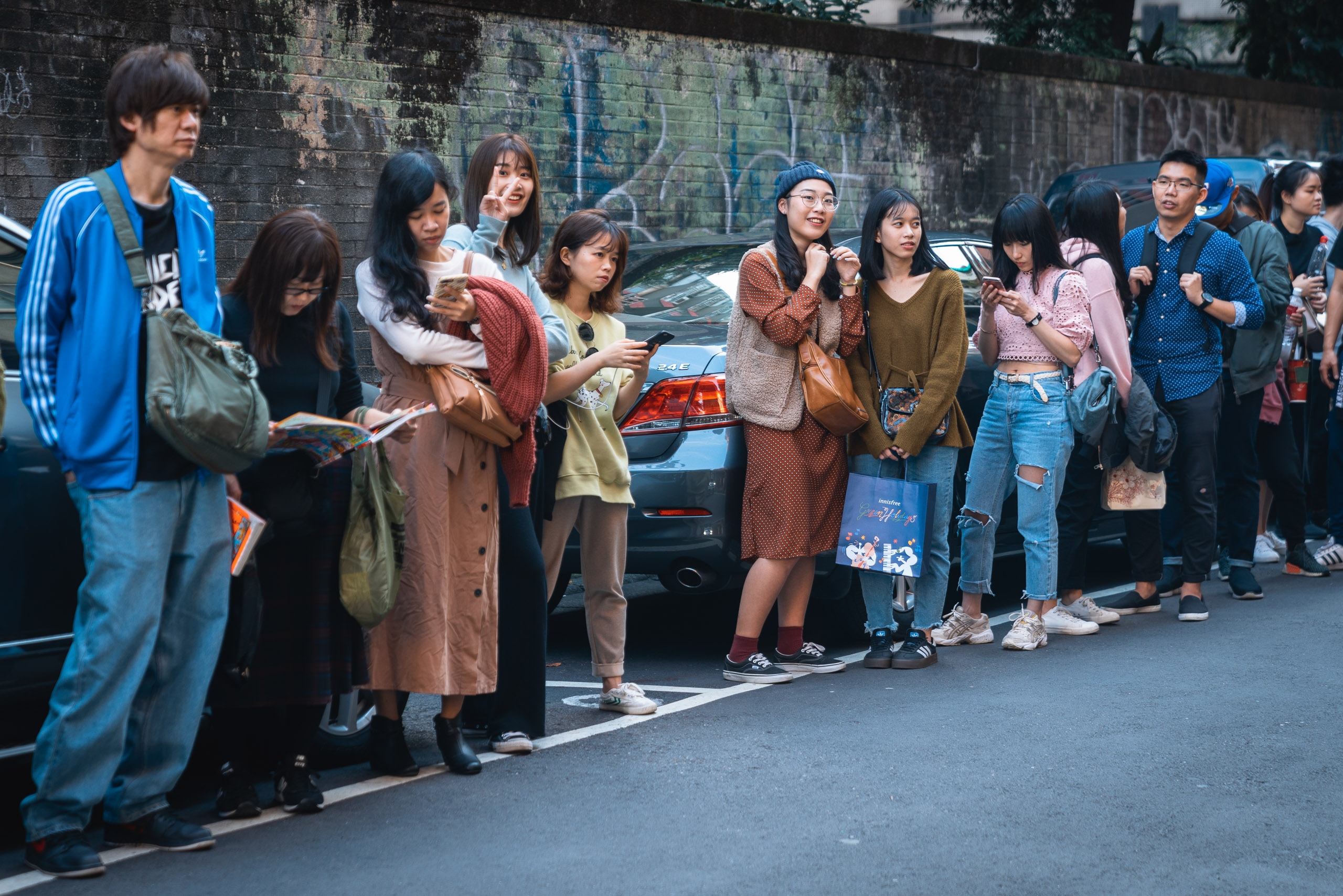 A group of young people stand in a line on an urban street against a graffitied brick wall, with parked cars behind them in what appears to be a casual gathering or event in Taipei
