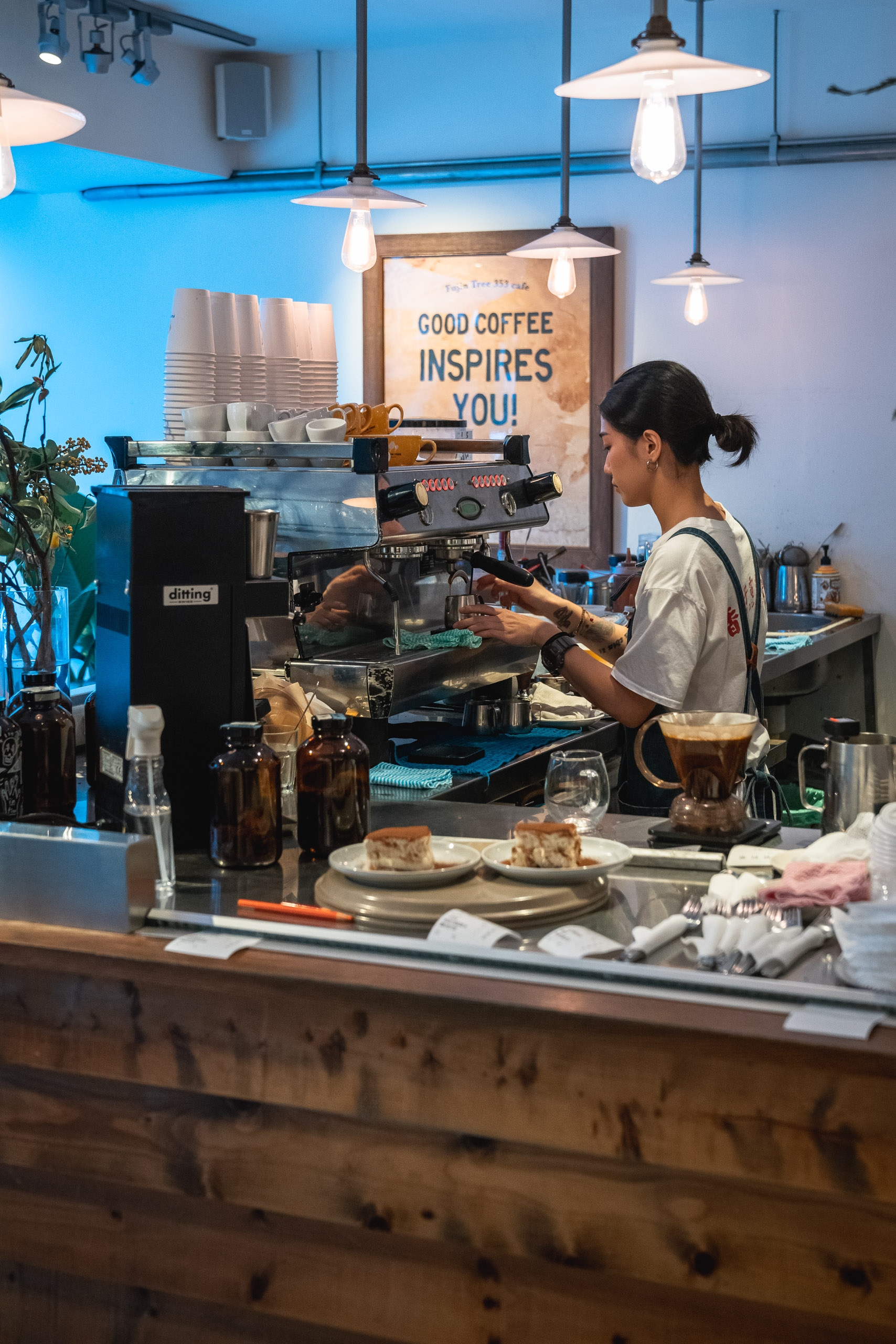 A barista operates an espresso machine behind a wooden counter in a modern café with blue accent lighting and a motivational sign reading "Good Coffee Inspires You"