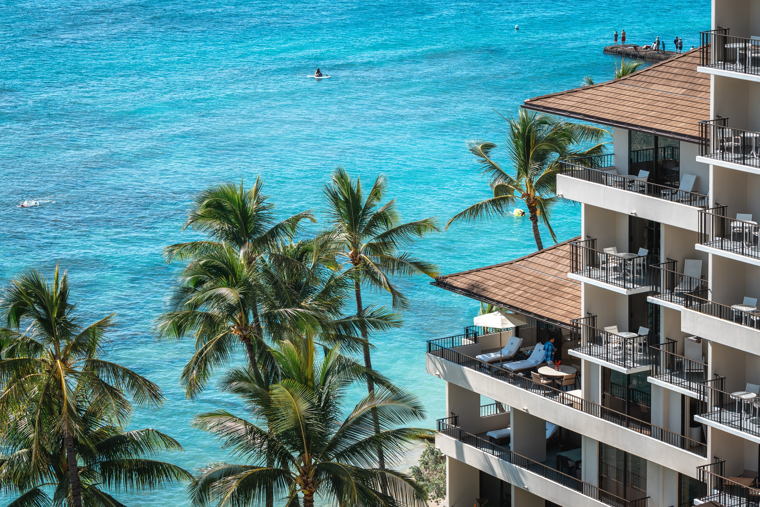 A multi-story beachfront hotel with white balconies overlooks turquoise ocean waters and swaying palm trees, with swimmers and a pier visible in the distance