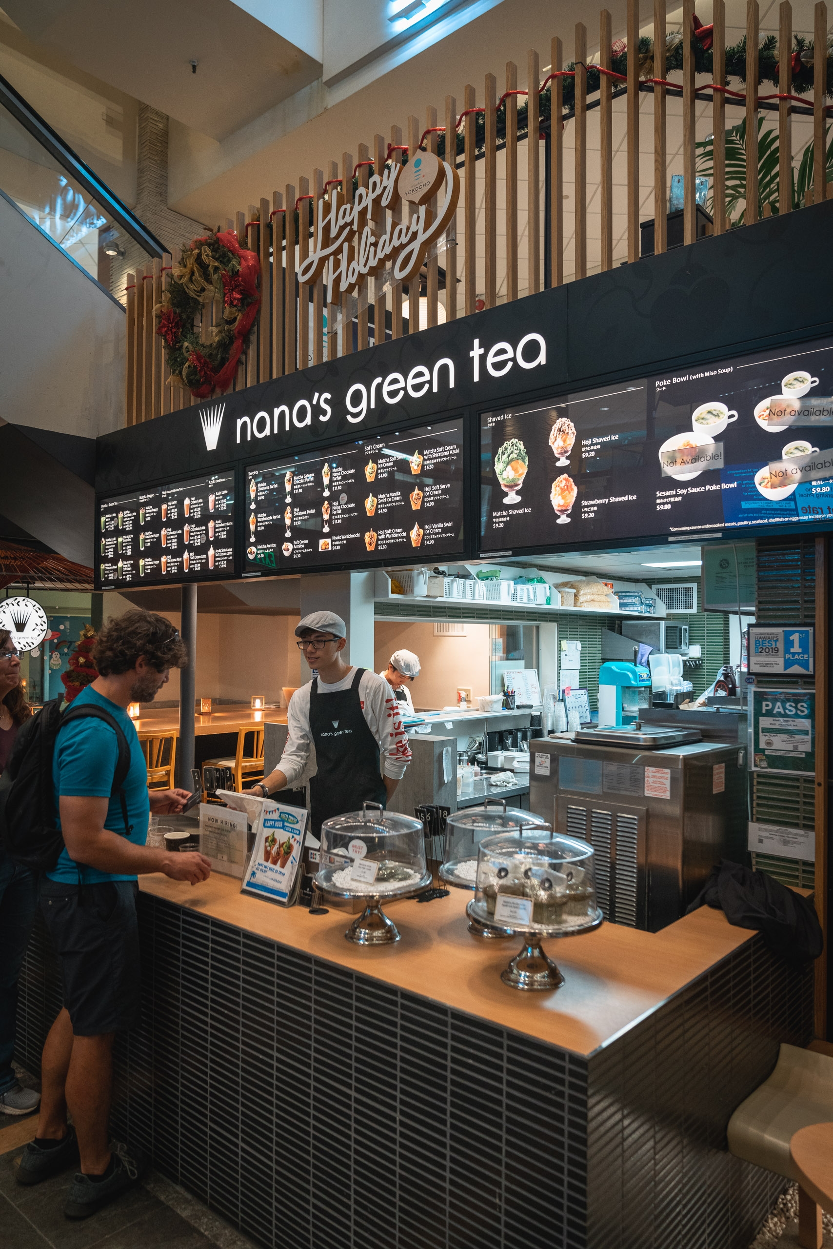 A customer orders at nana's green tea café counter in an indoor shopping mall, with menu boards above and display cases of pastries below, staff visible behind the counter