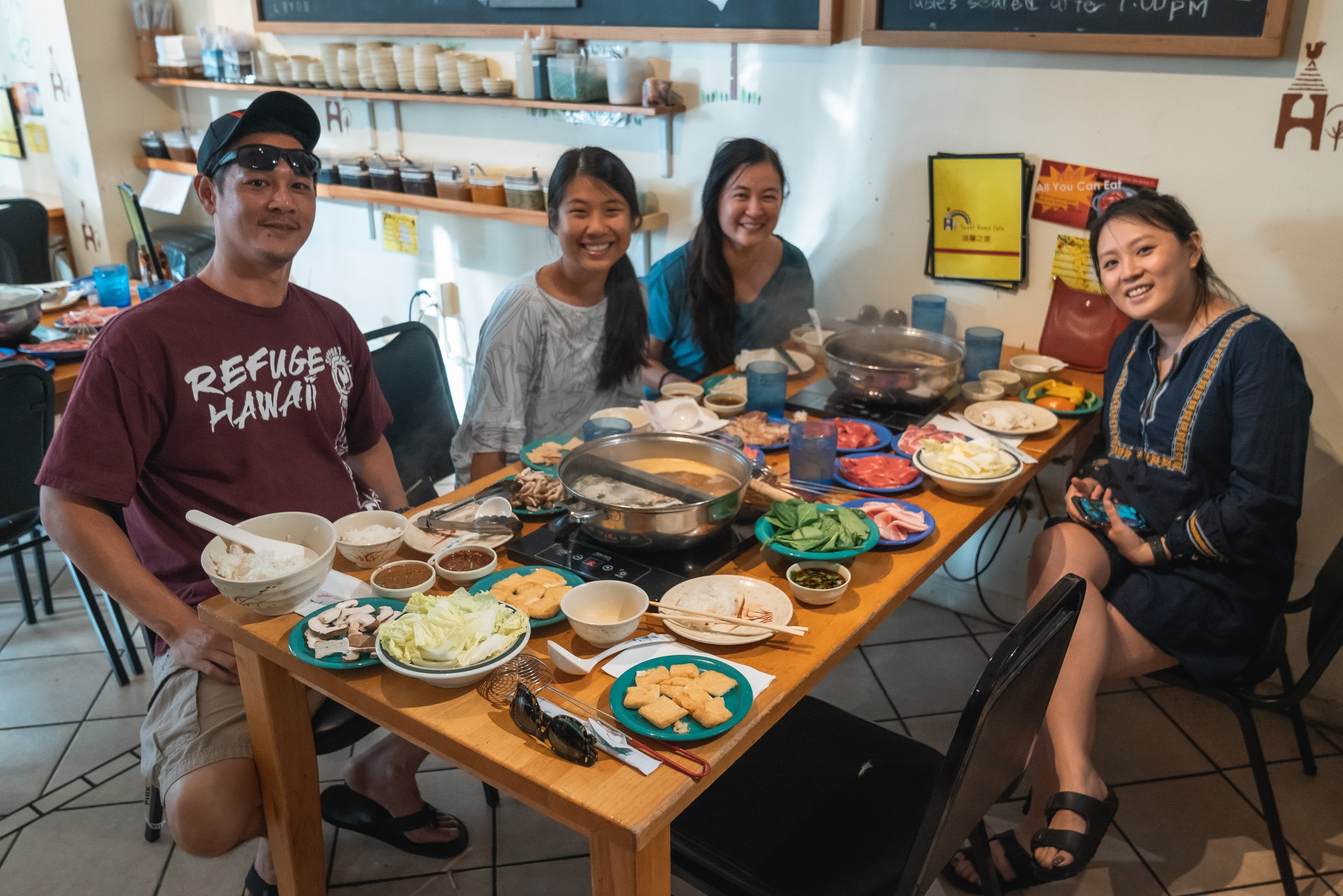 Four people smile at a wooden dining table laden with Asian dishes, hot pot, and condiments in a bright kitchen lined with shelves and chalkboard menu boards