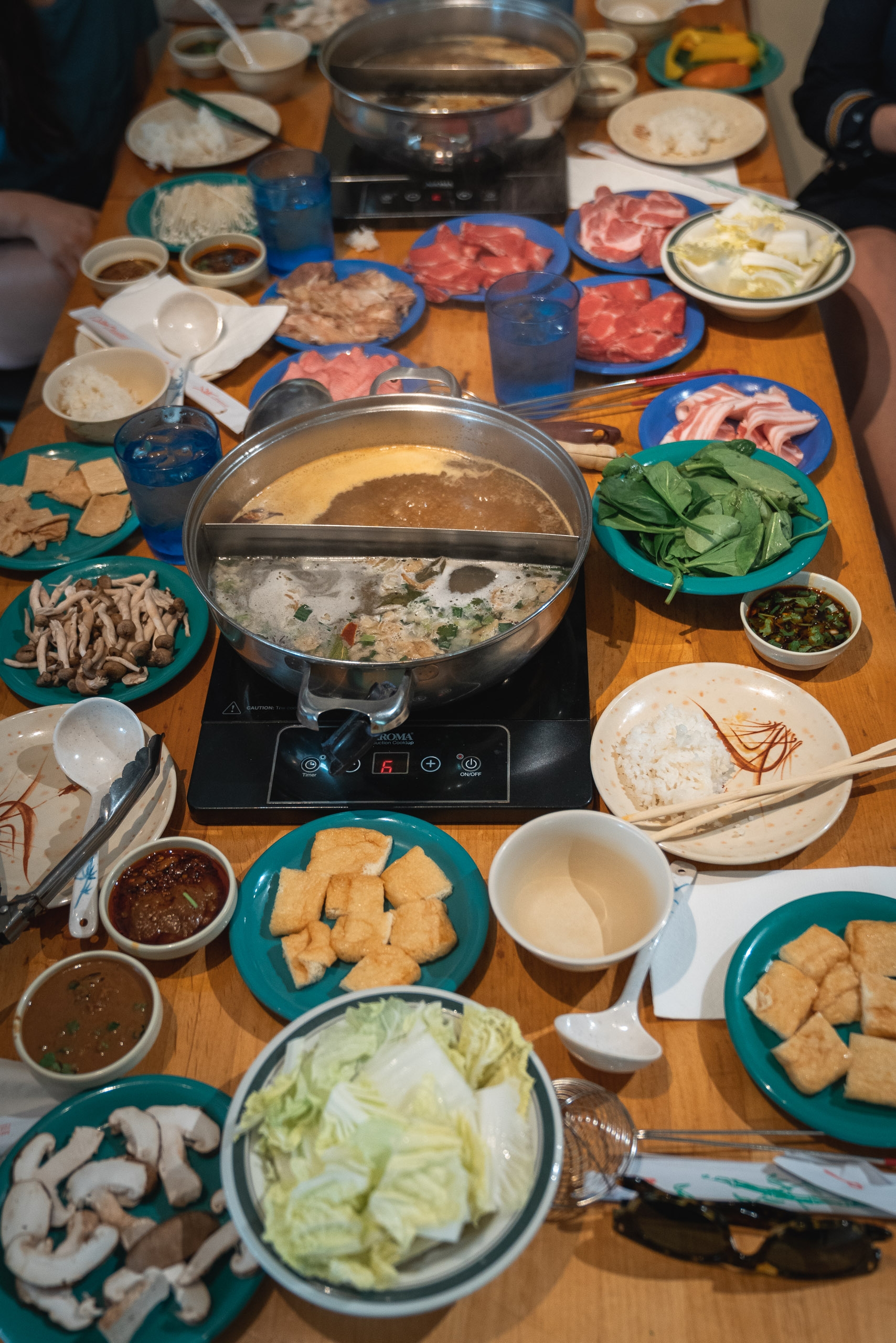 A hot pot meal setup with a divided broth pot at center, surrounded by plates of raw ingredients including thinly sliced meats, fresh vegetables, tofu, and noodles on a wooden table