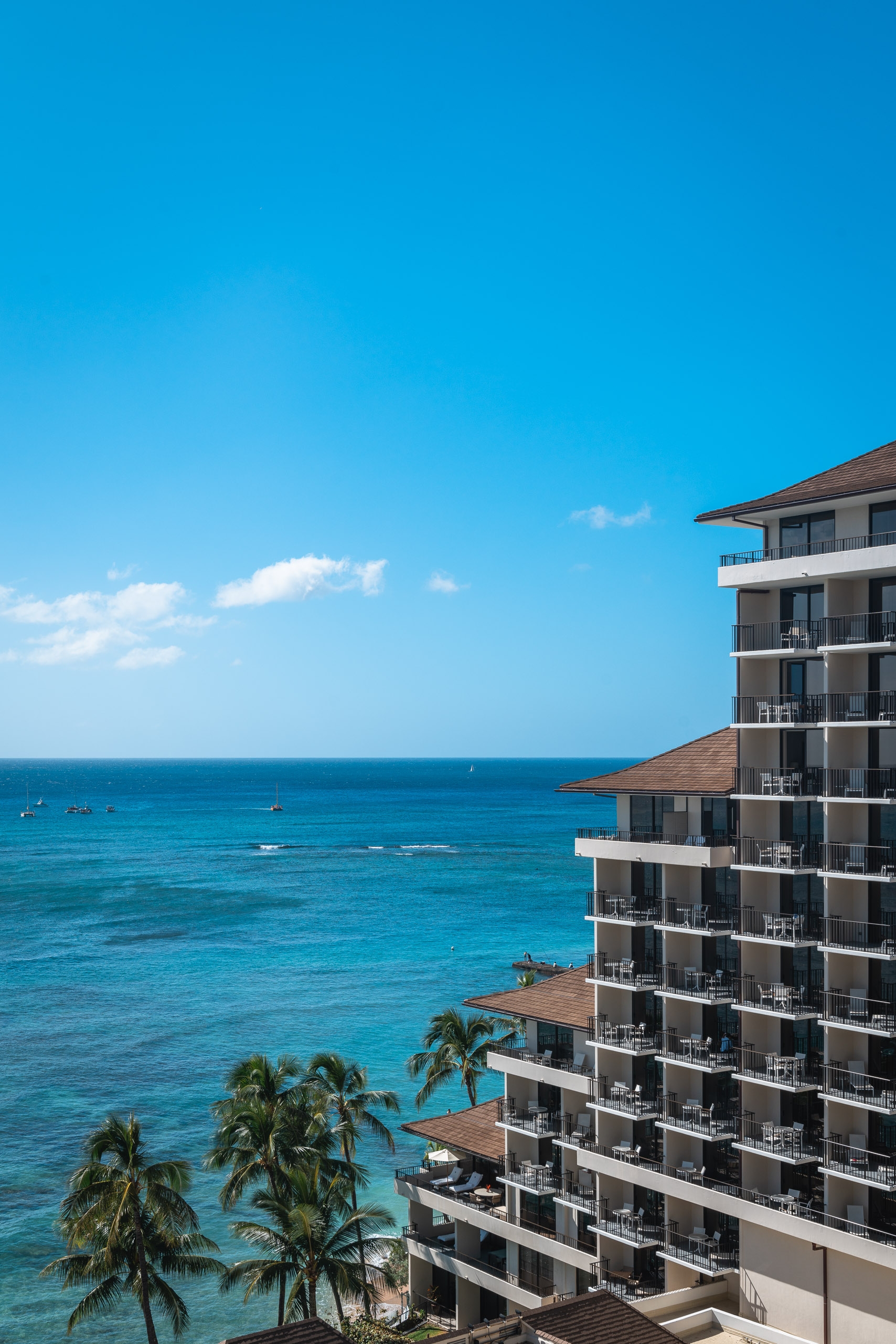 Modern high-rise hotel with tiered balconies overlooks turquoise ocean waters and palm trees under clear blue sky
