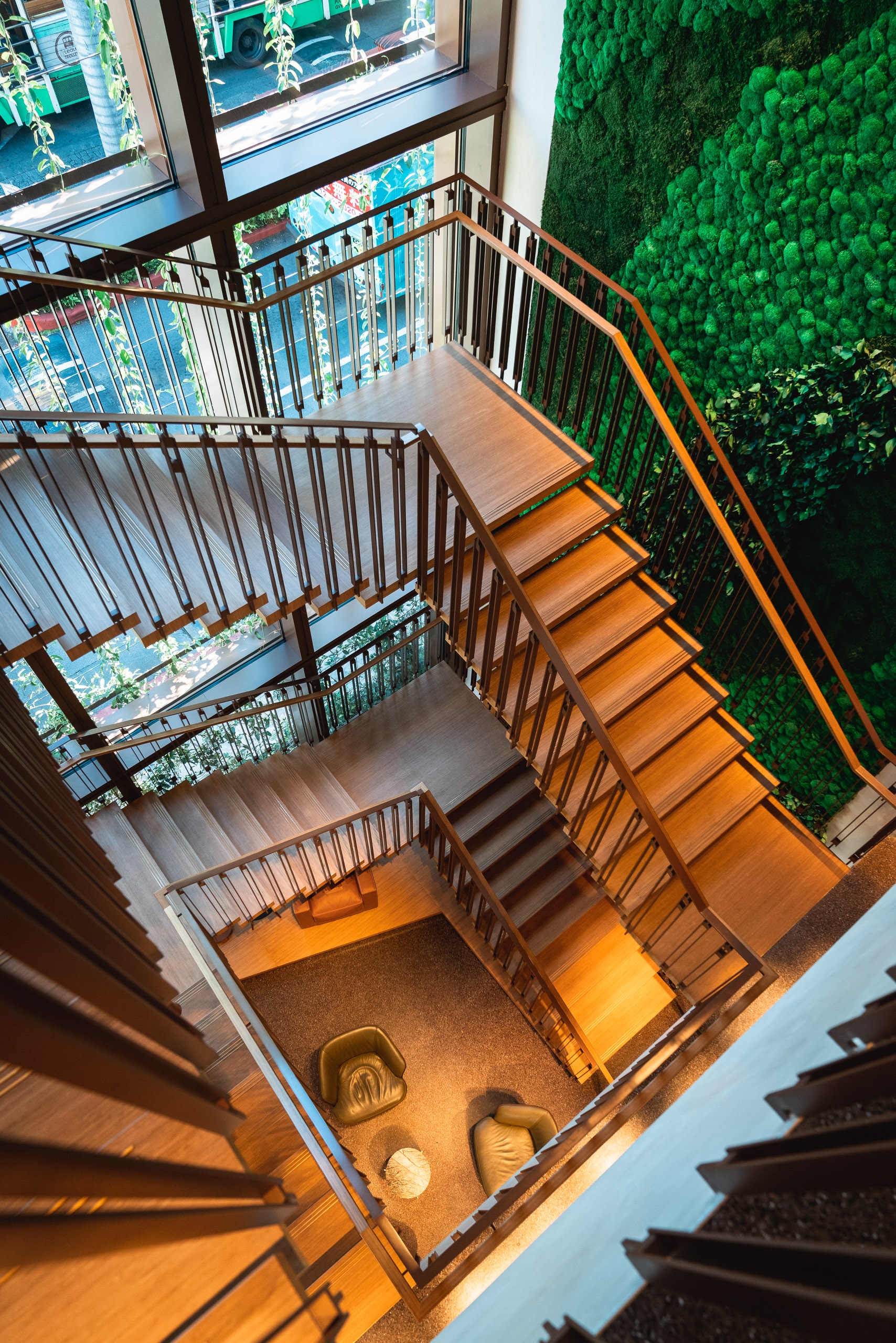 An overhead view of a modern stairwell with wooden steps and metal railings, flanked by a vibrant green moss wall and windows overlooking a bright urban street