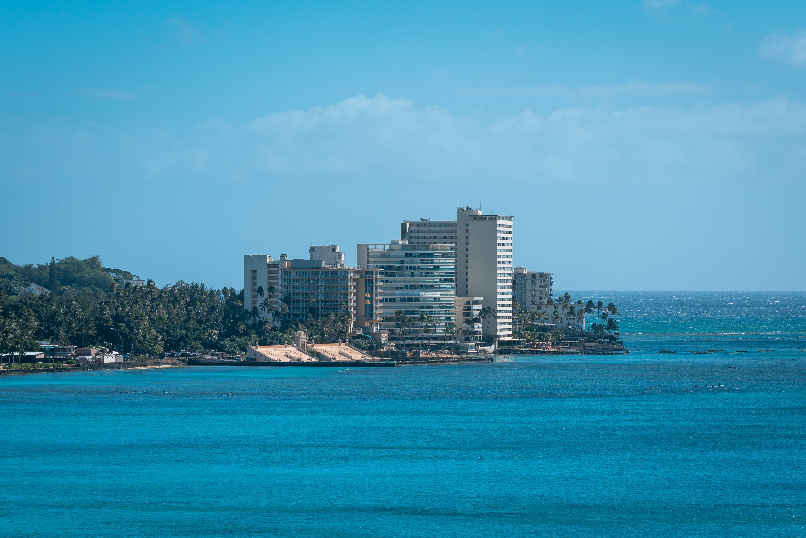 Modern high-rise hotels line a sandy beach on Oahu's coast, surrounded by lush green vegetation beneath clear blue skies and turquoise ocean waters