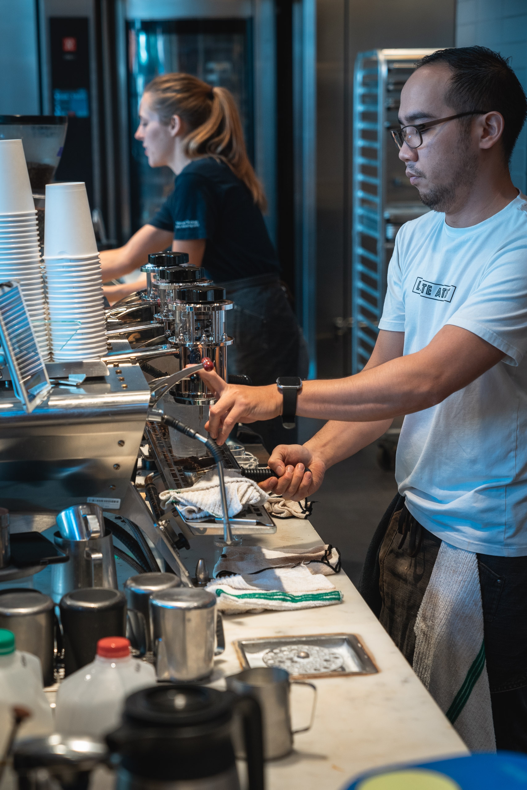 A man in glasses operates a professional espresso machine while a woman arranges cups nearby in a bright, modern blue-lit coffee shop or training facility