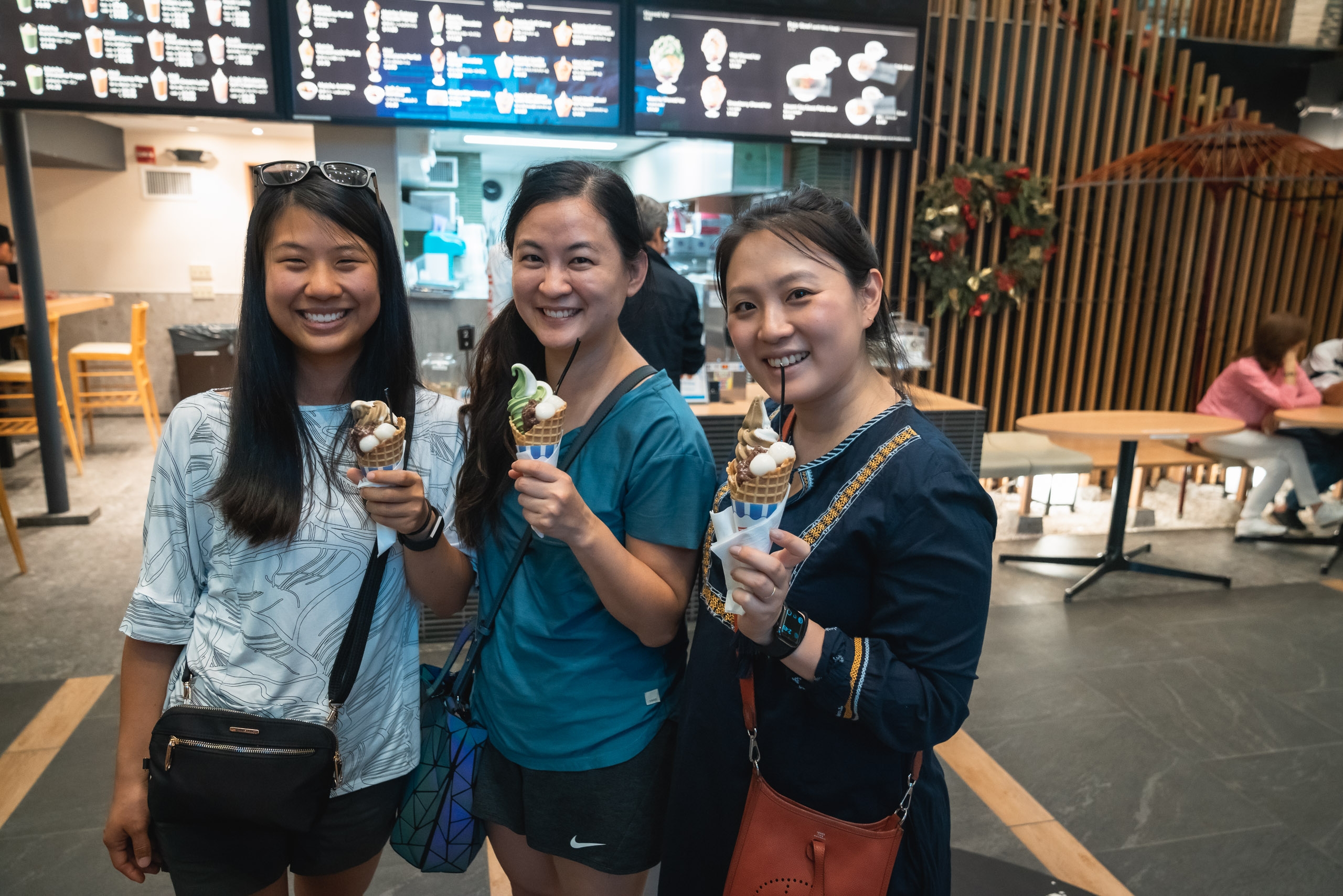 Three smiling women hold ice cream cones inside a modern café with illuminated menu boards and wooden interior elements visible behind them
