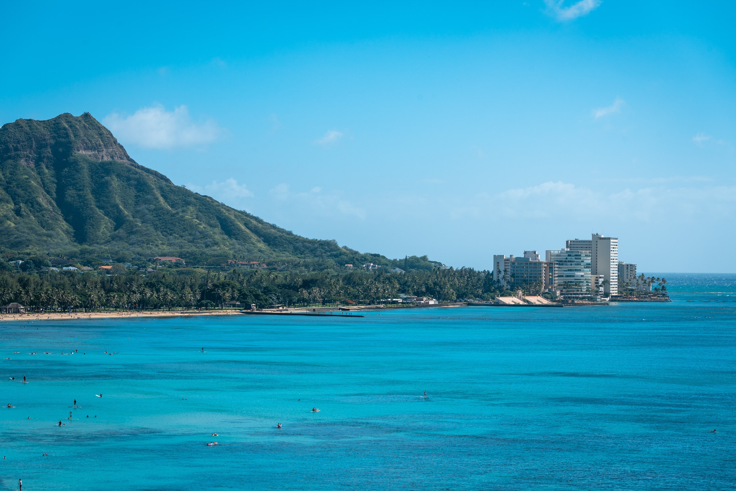 Turquoise waters dotted with swimmers stretch toward a dramatic green volcanic mountain rising behind beachfront hotels and palm trees under clear blue sky