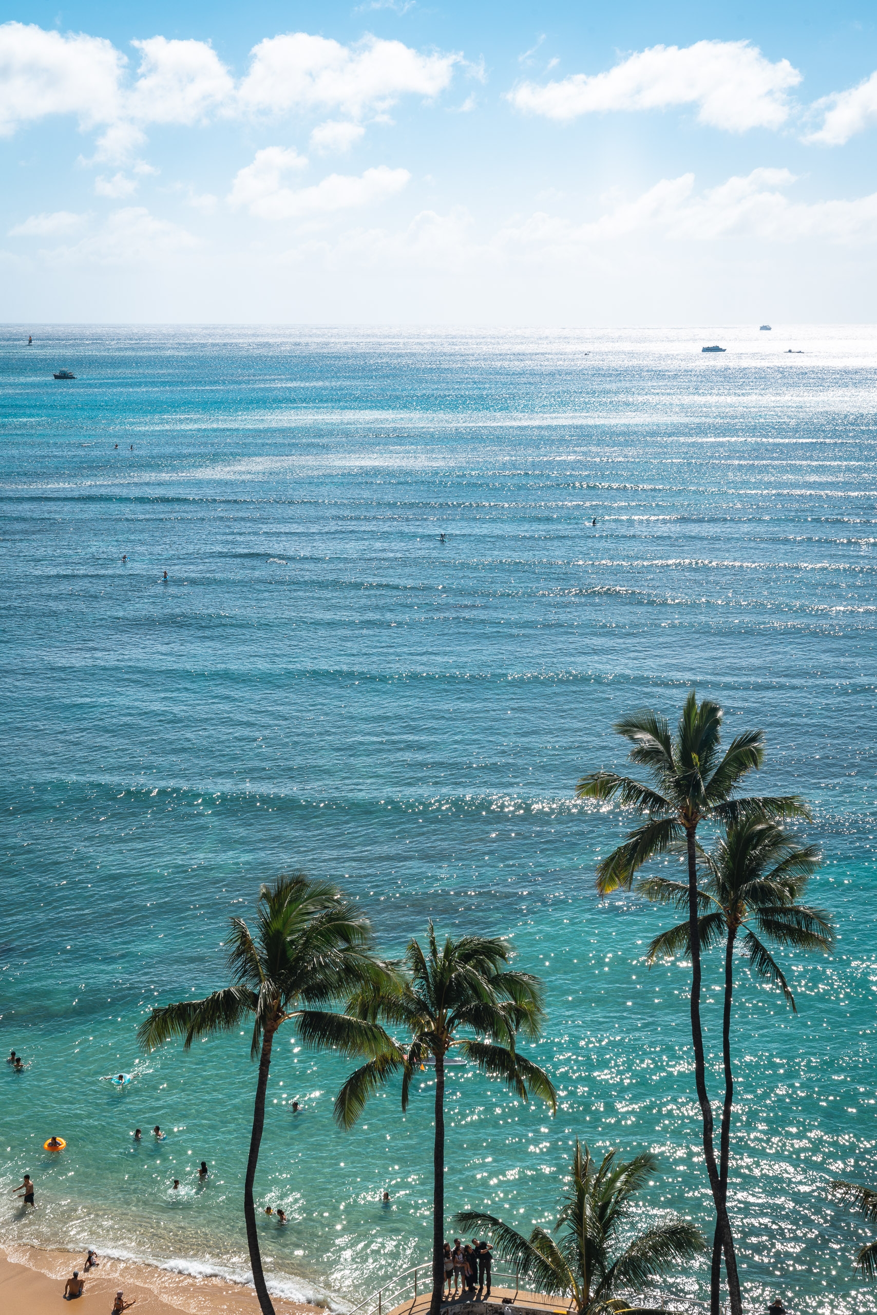 Tropical beach scene with palm trees framing turquoise waters, swimmers dotting the sandy shore, and bright sunlight reflecting across the calm ocean under a partly cloudy sky