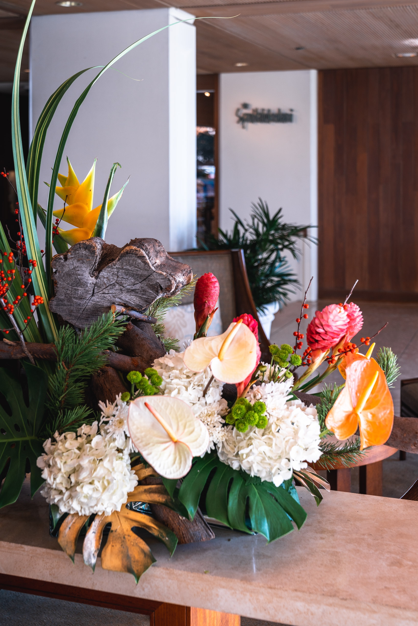 An elaborate floral arrangement sits on a reception desk, featuring white hydrangeas, tropical anthuriums in yellow and orange, red ginger flowers, and lush green foliage arranged in a textured dark container against a modern hotel lobby backdrop