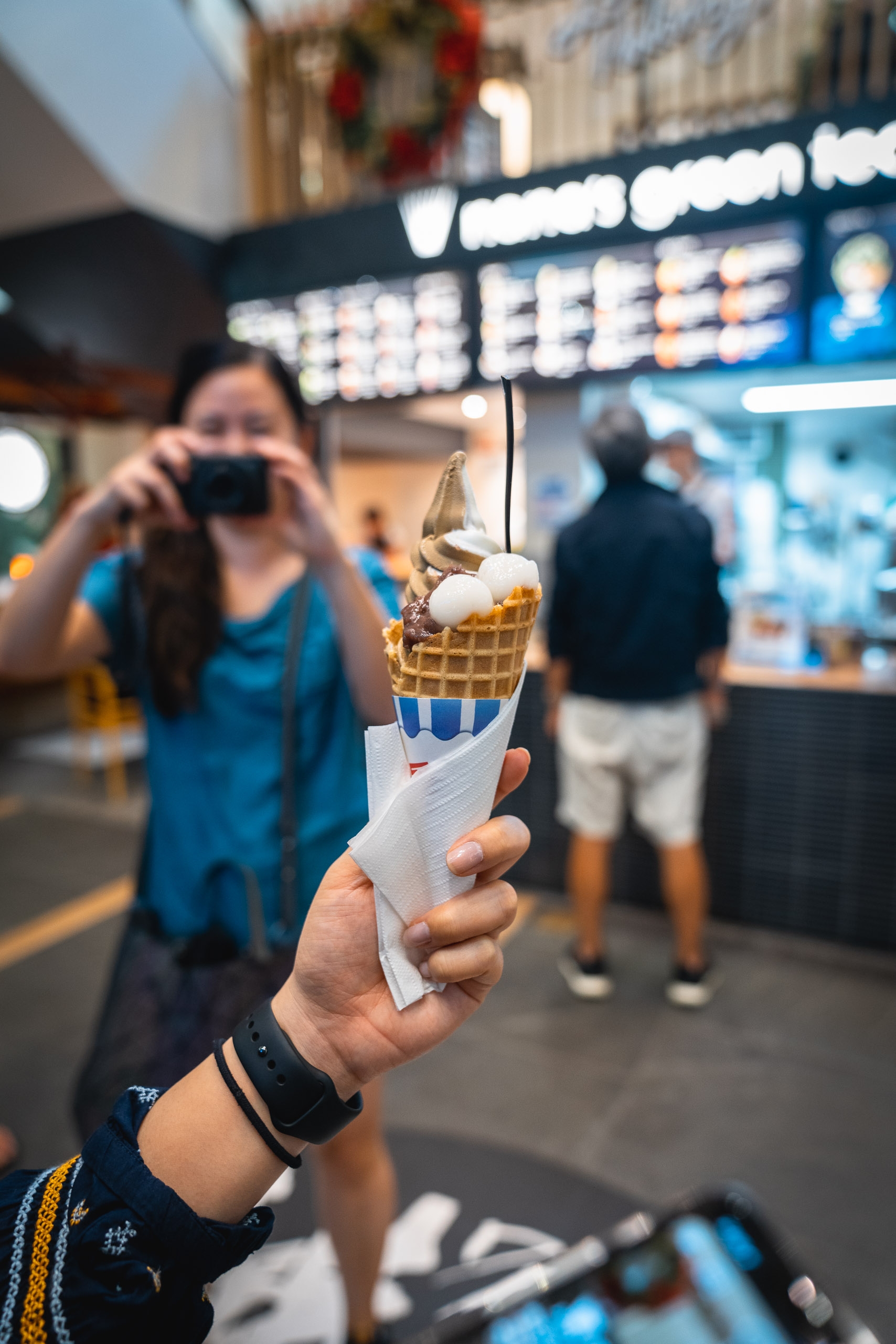 A hand holds a vanilla ice cream cone wrapped in paper at a beachside shop, with a blurred woman photographing it and storefront signage visible in the background.