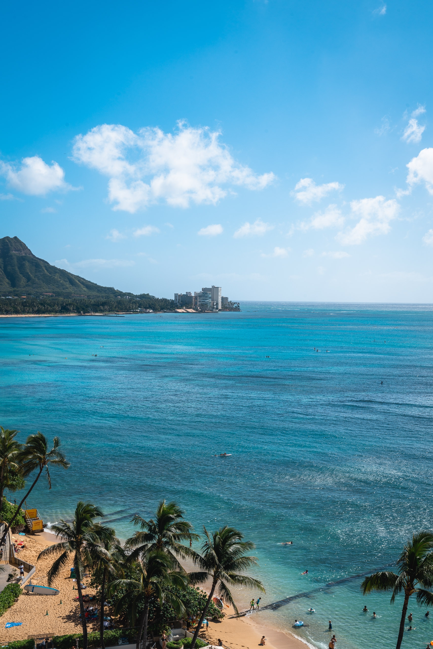 Sandy Waikiki Beach curves beneath a verdant volcanic mountain, with turquoise waters, palm trees, and high-rise hotels visible across the clear blue Pacific horizon