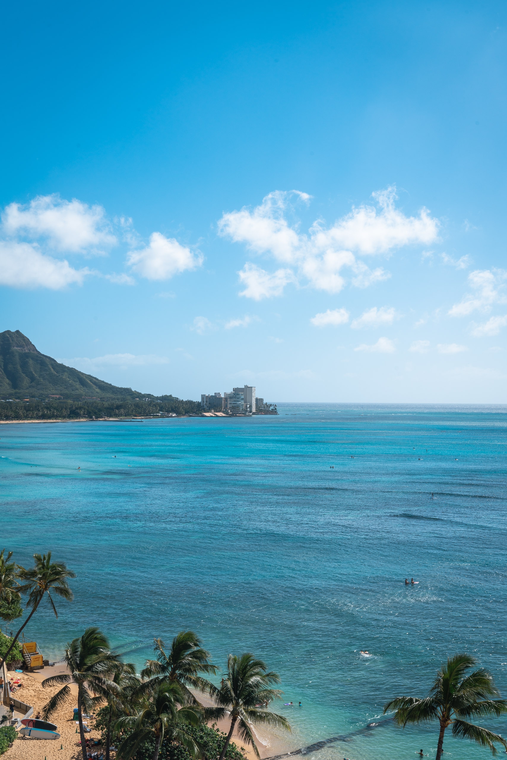 Turquoise waters and white sandy beach frame Waikiki's shoreline, with Diamond Head volcano rising dramatically on the left and high-rise hotels visible across the bay under bright blue skies