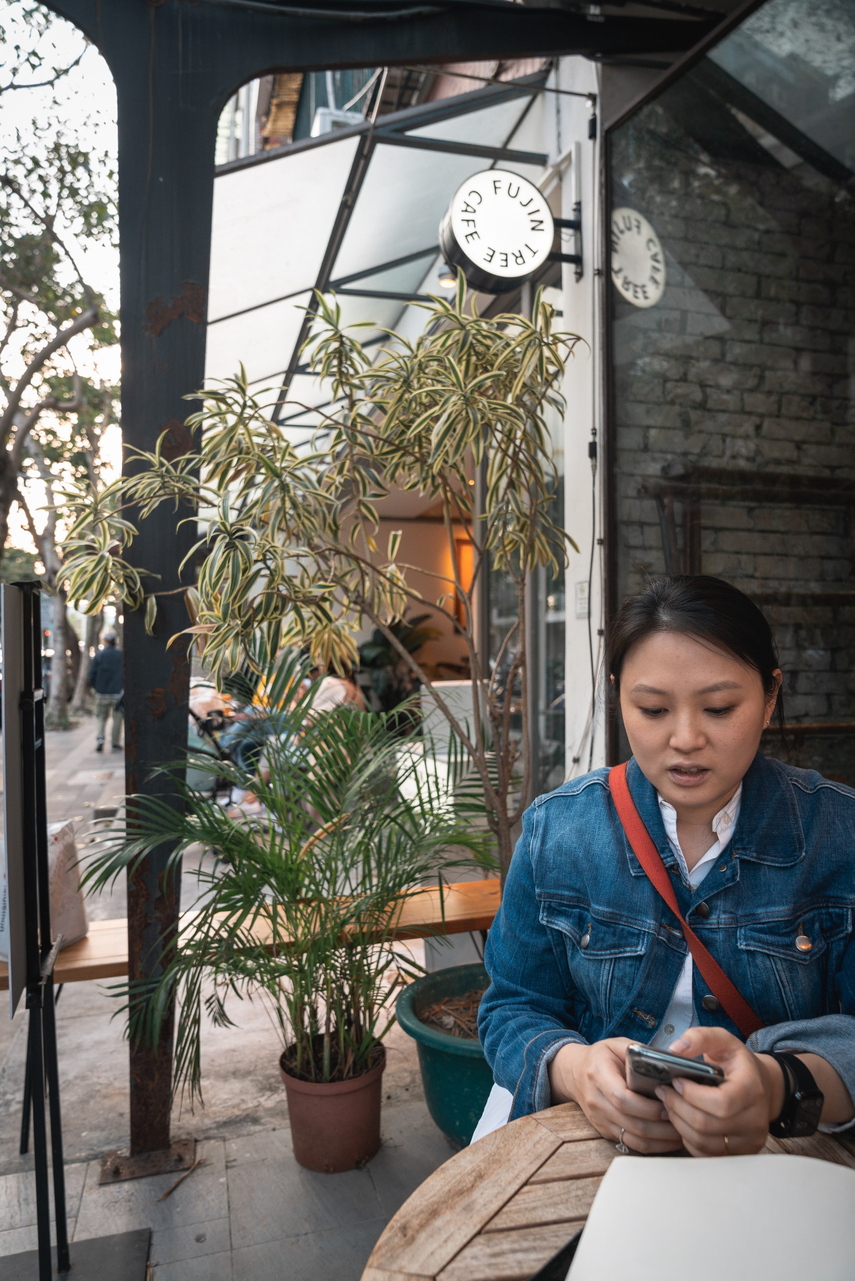 A woman in a denim jacket sits at an outdoor café table, focused on her phone, surrounded by hanging plants and metal structural beams beneath a covered storefront