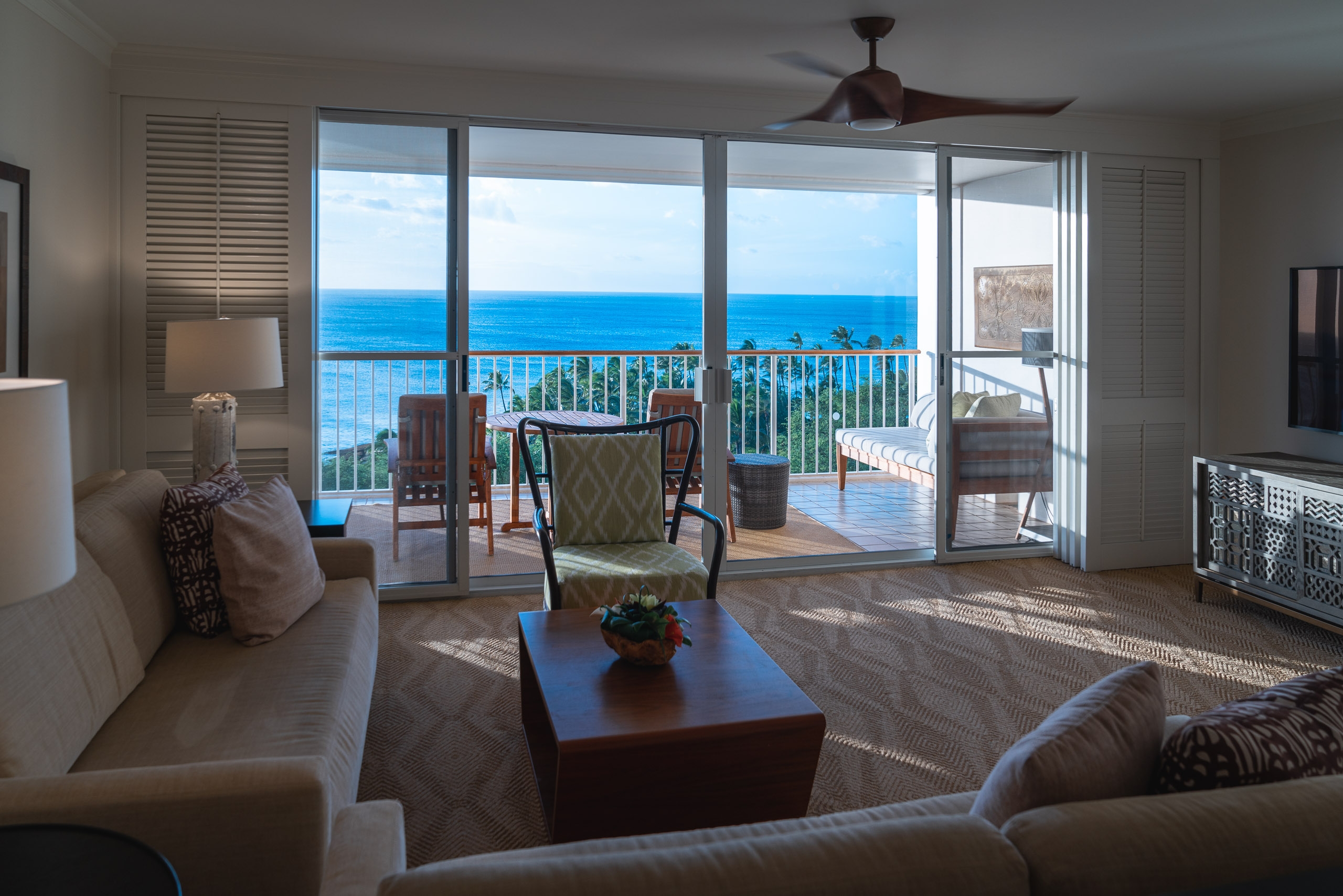 A spacious beachfront resort suite features modern furnishings, expansive glass doors opening to a balcony overlooking turquoise ocean waters, and bright natural daylight streaming across the neutral-toned interior