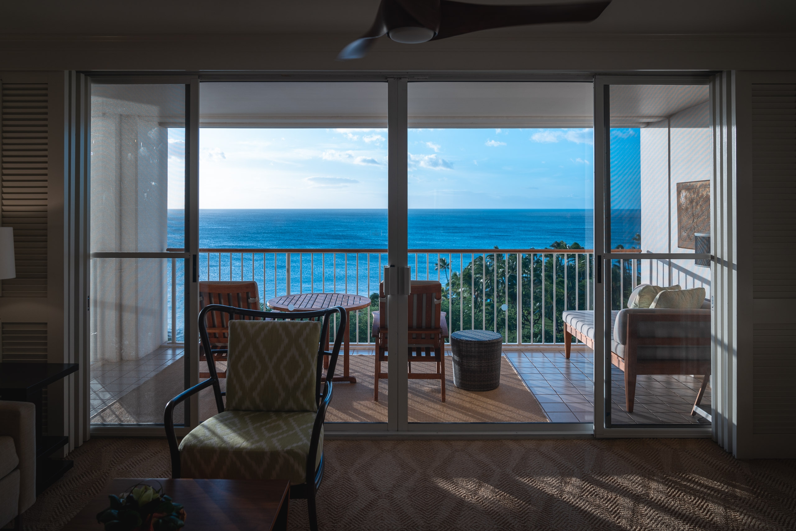 Large glass doors open onto a beachfront terrace with lounge chairs overlooking the bright blue Pacific Ocean and distant horizon under clear sky